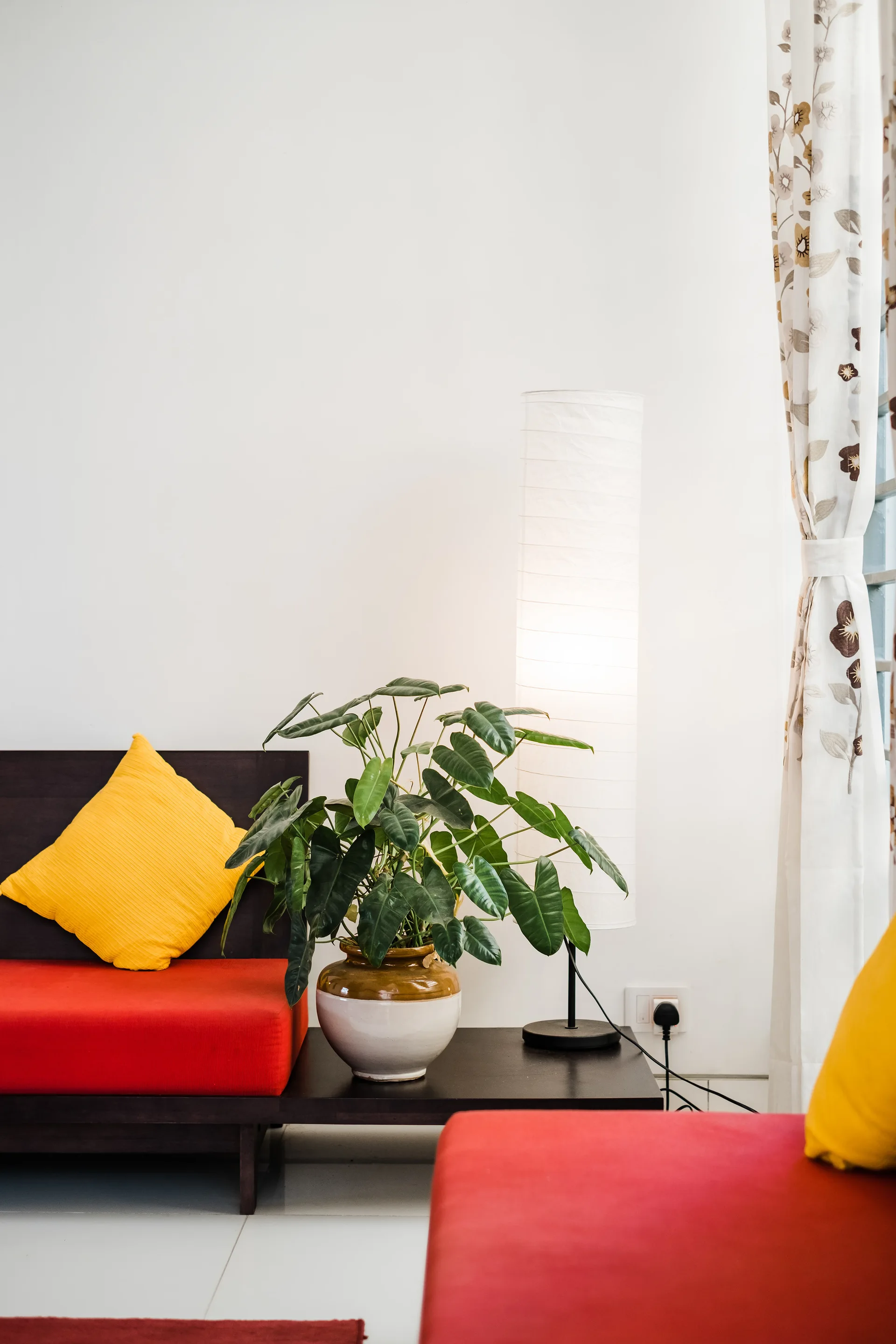 Red upholstered seating with yellow pillows is arranged next to a dark wood side table holding a leafy green plant and a glowing paper floor lamp.