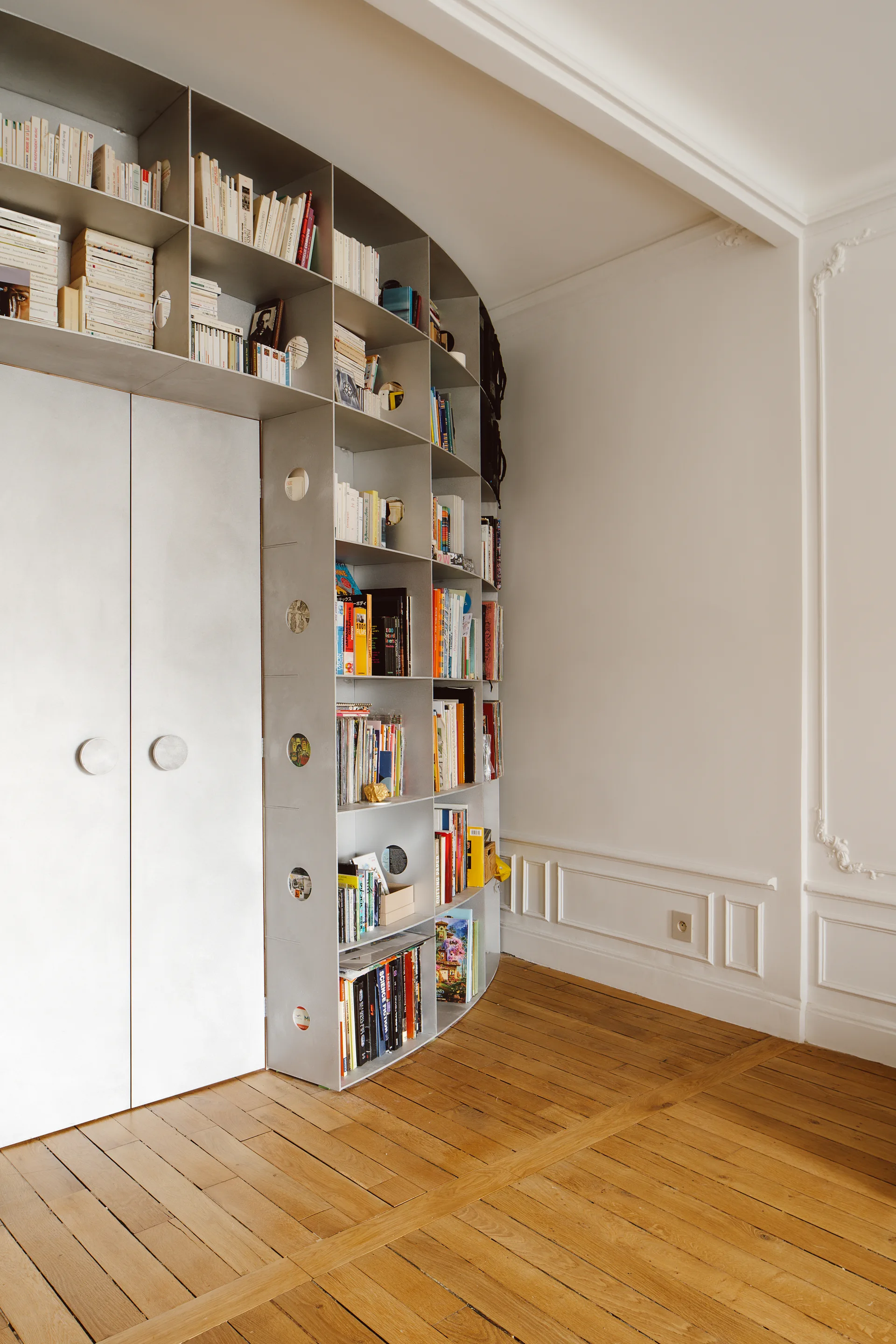 A curved light gray metal bookshelf with circular cutouts stands on warm parquet wood flooring next to traditional white wall paneling.