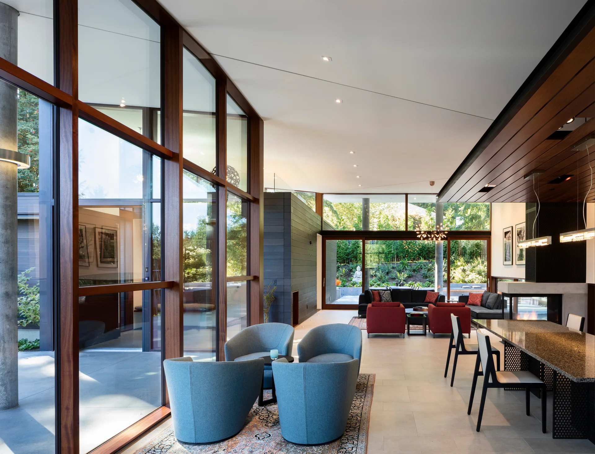 A spacious, open-plan living room features tall wood-framed glass walls, blue armchairs on a patterned rug, and a modern dining table by a fireplace.