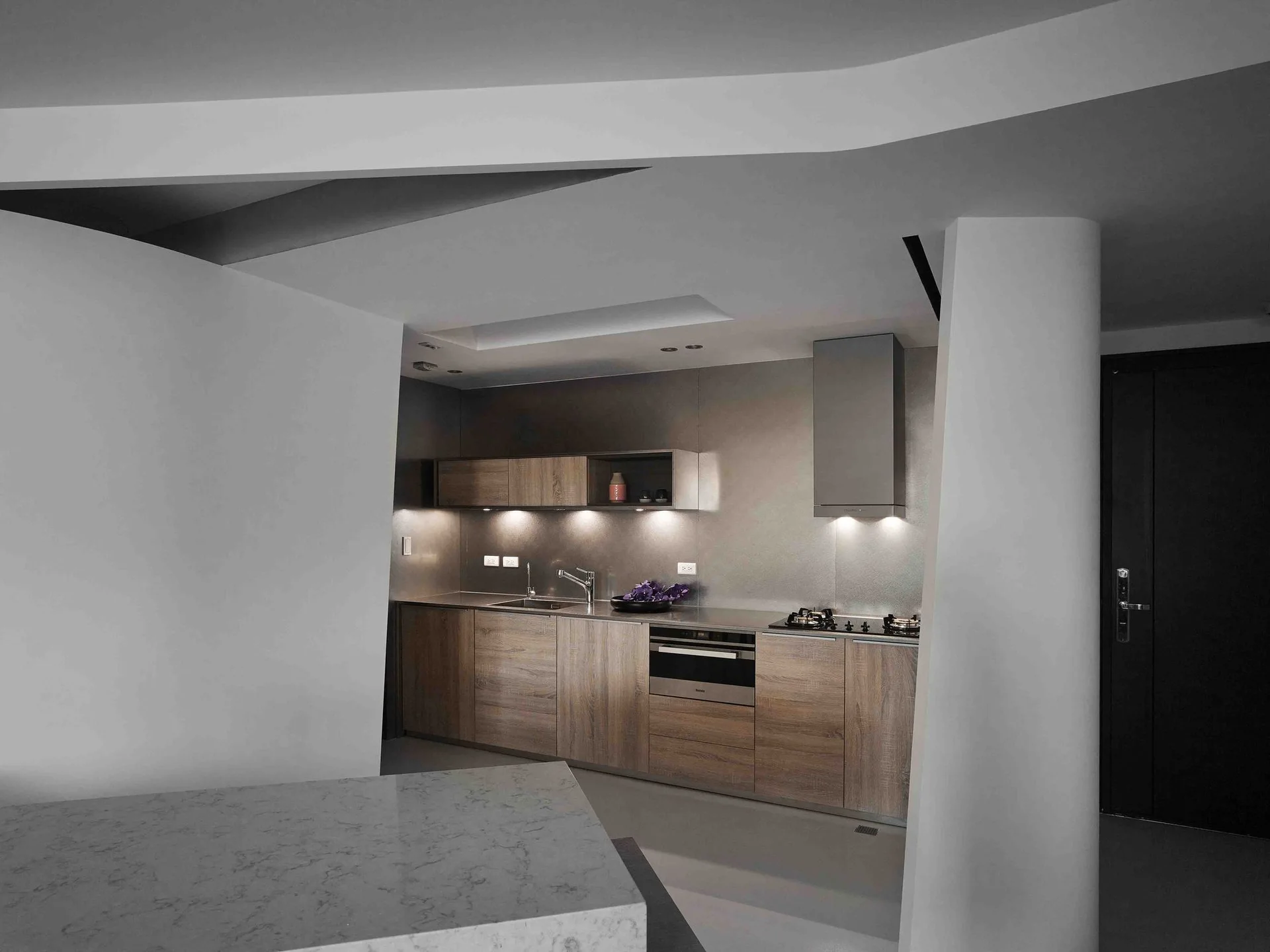 A minimalist kitchen features flat-panel wood cabinets, a stainless steel range hood, and a prominent curved white structural column and ceiling.