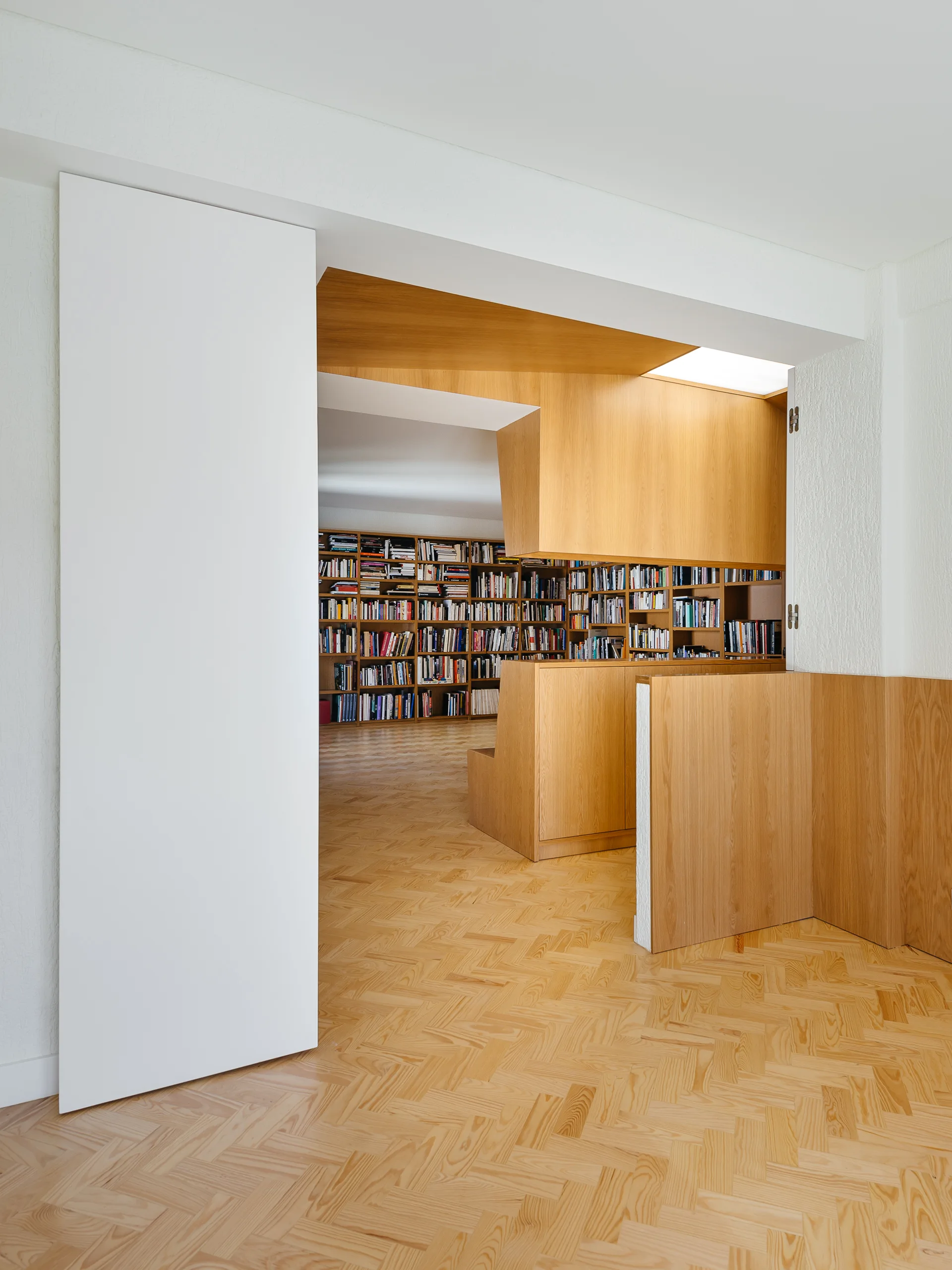 A view through a white doorway shows a minimalist home library with extensive custom wood bookshelves and a herringbone parquet wood floor.