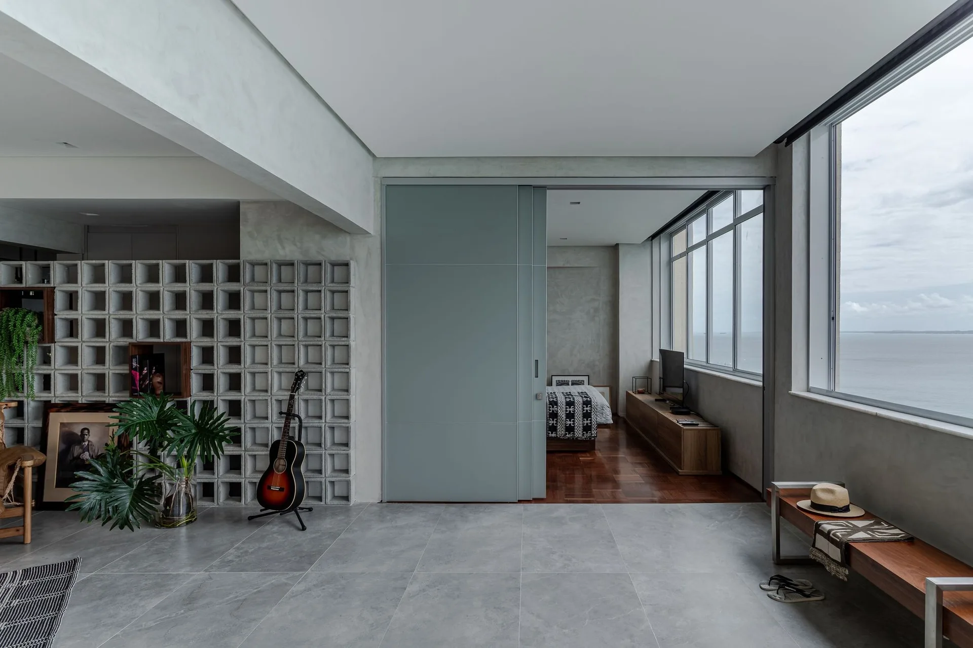A modern grey living room features a patterned concrete block screen, a large sliding door, and a wooden bench with views of the ocean.