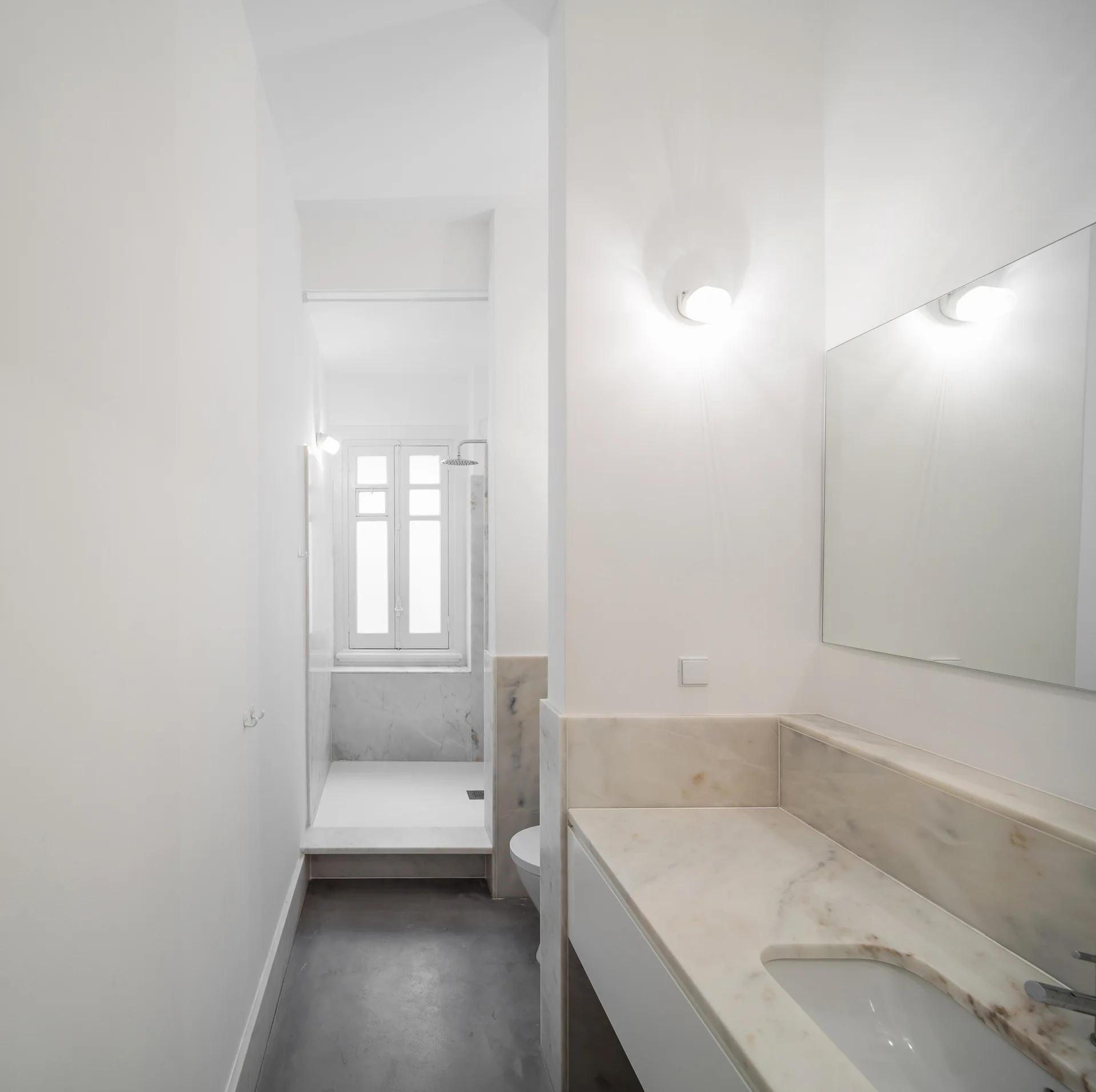 A narrow minimalist bathroom with a cream-veined marble vanity, a large mirror, grey concrete flooring, and a walk-in shower with a window.