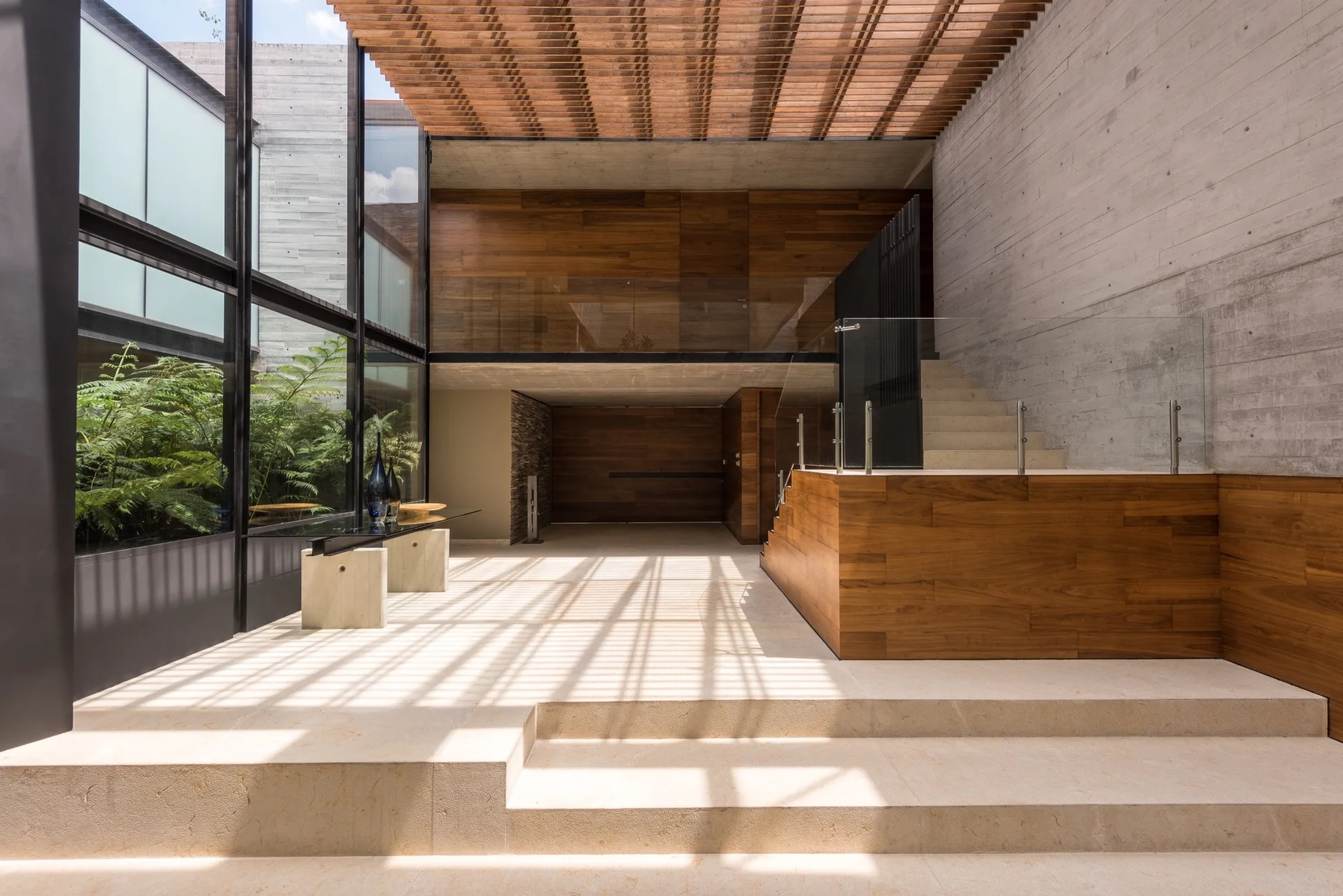 A modern entryway features a slatted wood ceiling, board-formed concrete walls, stone tile floors, and a wooden staircase with a glass railing.