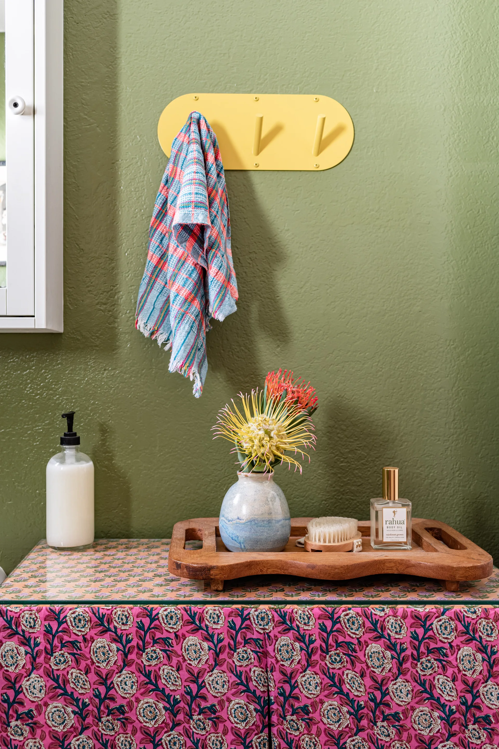 A pink floral patterned vanity cabinet is set against an olive green bathroom wall featuring a yellow wall hook with a colorful plaid towel.