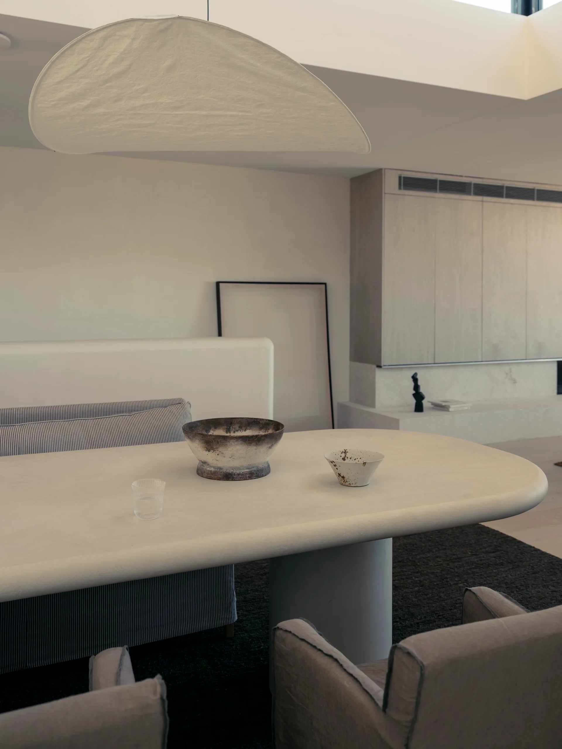 A minimalist dining room featuring a light-colored sculptural table with ceramic bowls, upholstered chairs, and a large paper pendant light overhead.