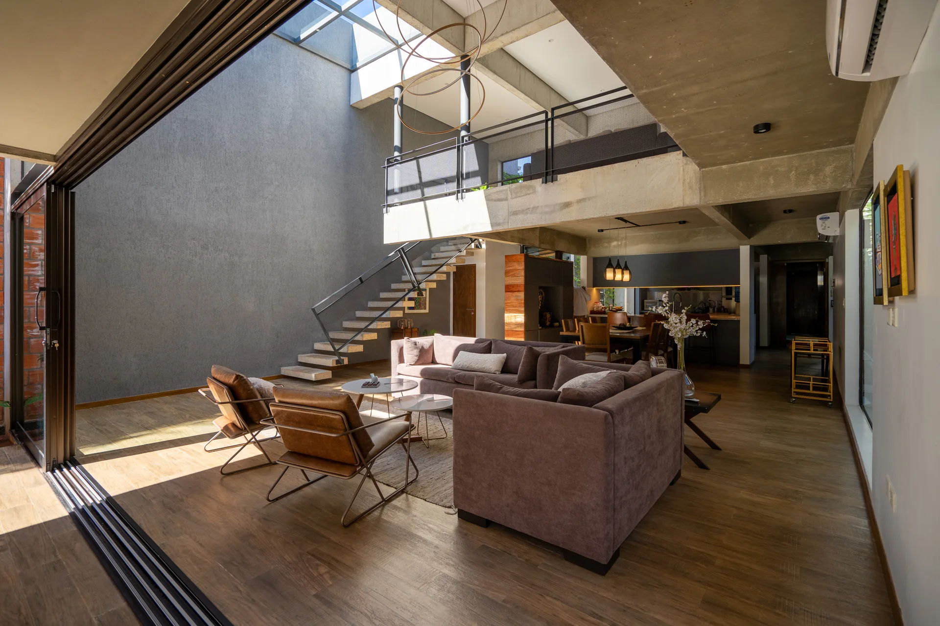 An open-plan living room featuring a brown sectional sofa, leather lounge chairs, an exposed concrete ceiling, and a floating staircase beneath a skylight.