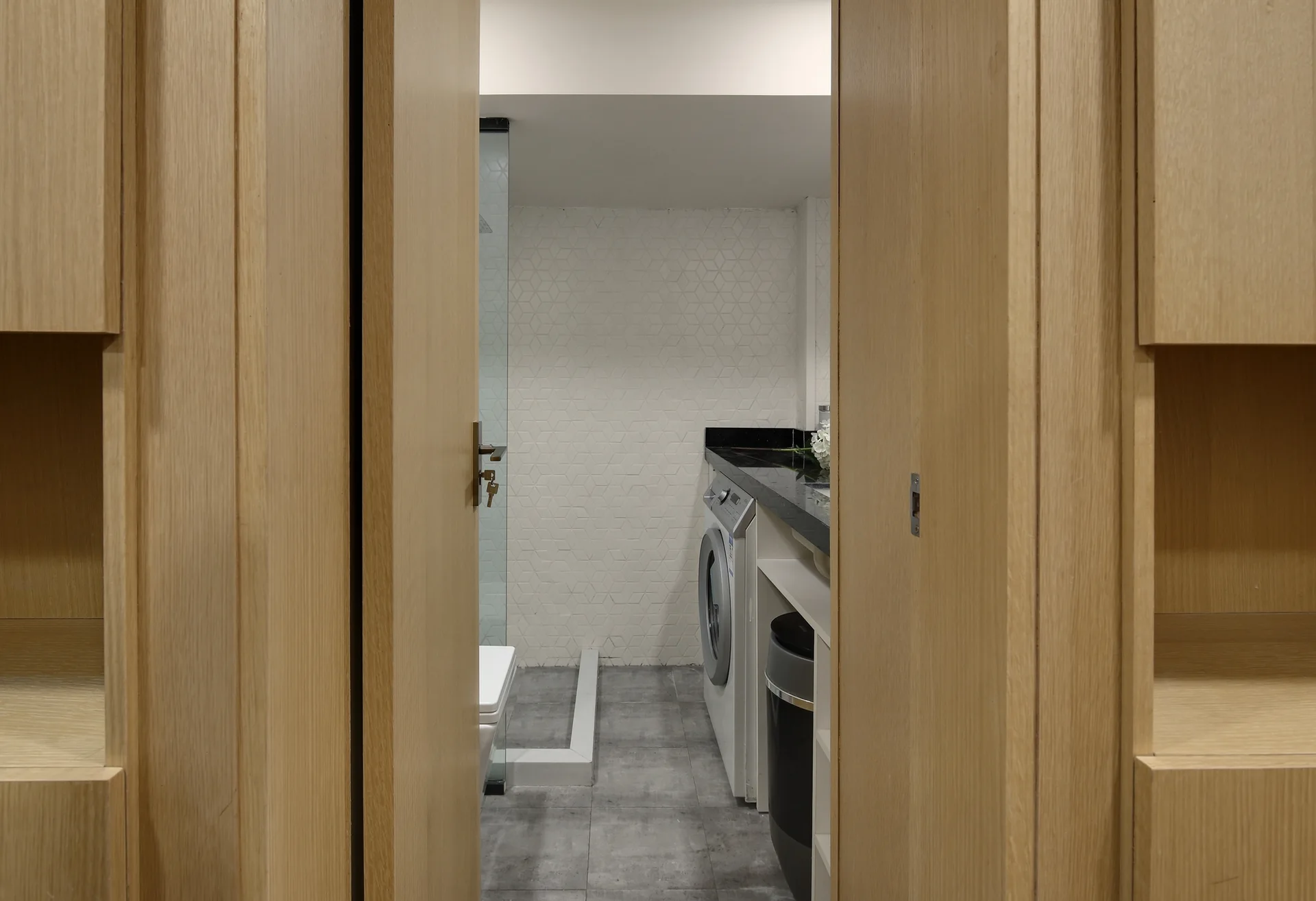 An open wooden doorway reveals a compact laundry room featuring a front-load washer under a black countertop with white geometric wall tiles.