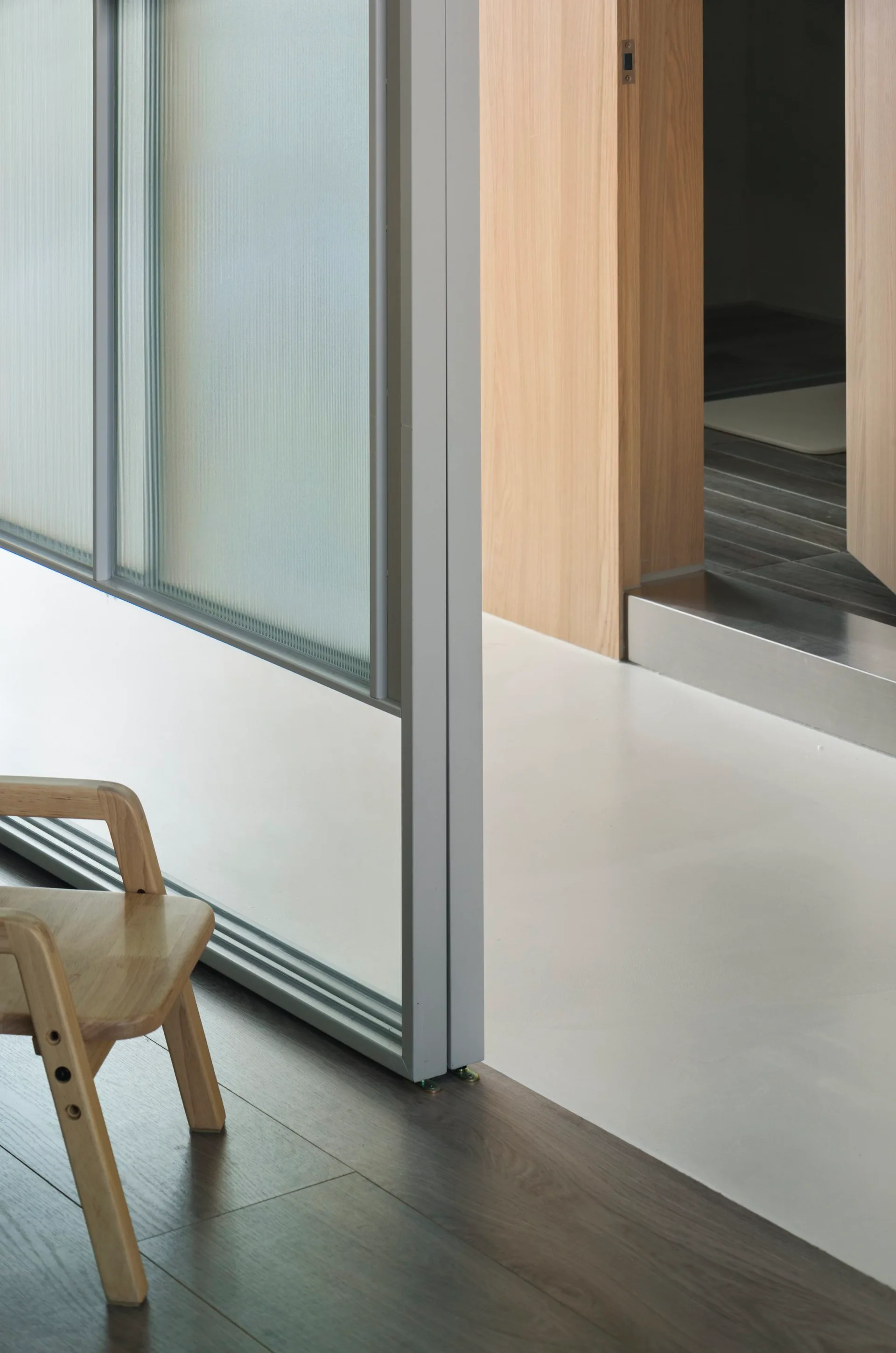 A wooden chair sits on dark wood floors next to a metal and frosted glass partition transitioning into a white-floored hallway.