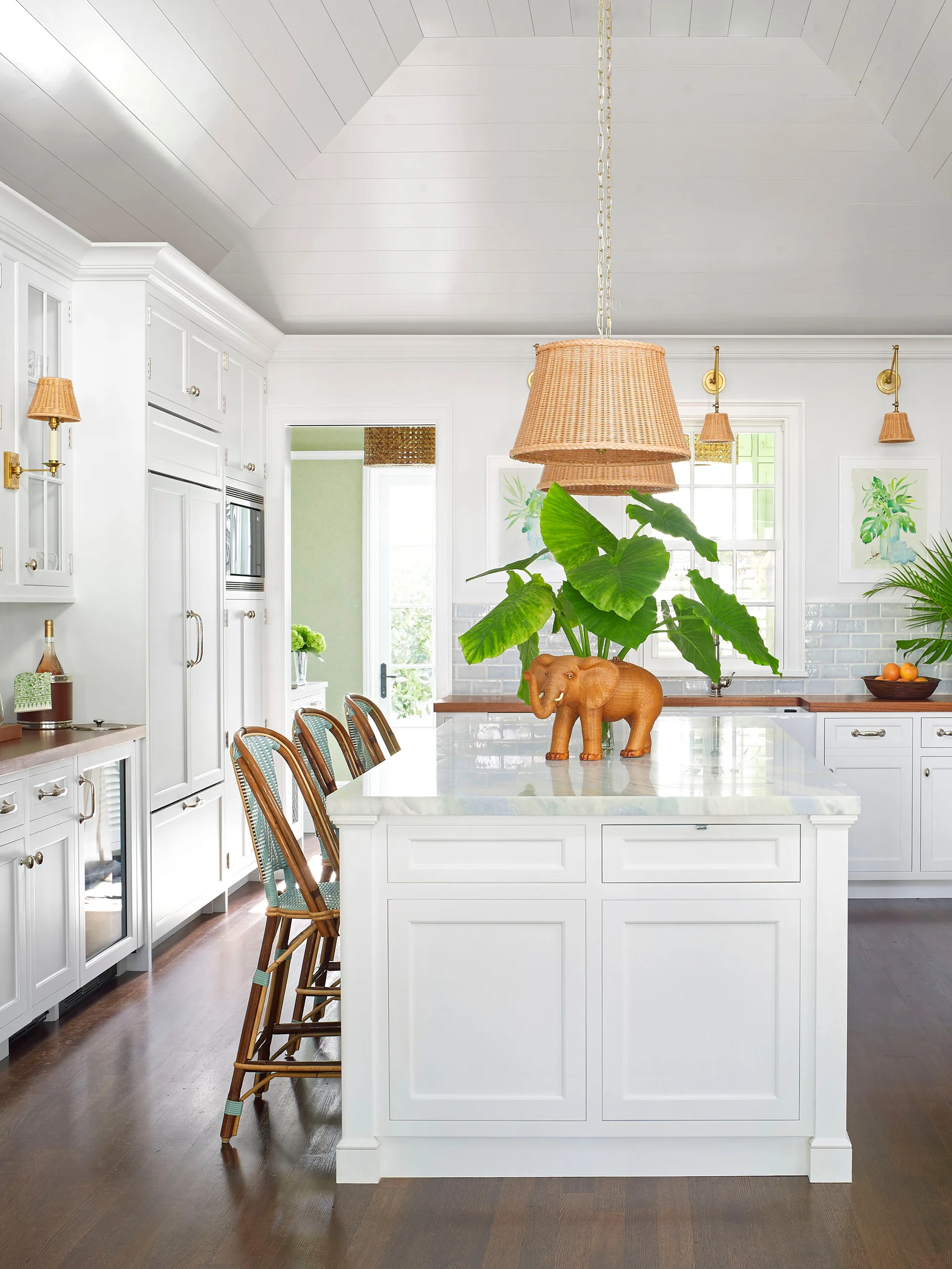 A bright white kitchen features a marble-topped island with woven barstools, a blue tile backsplash, and a vaulted paneled ceiling.