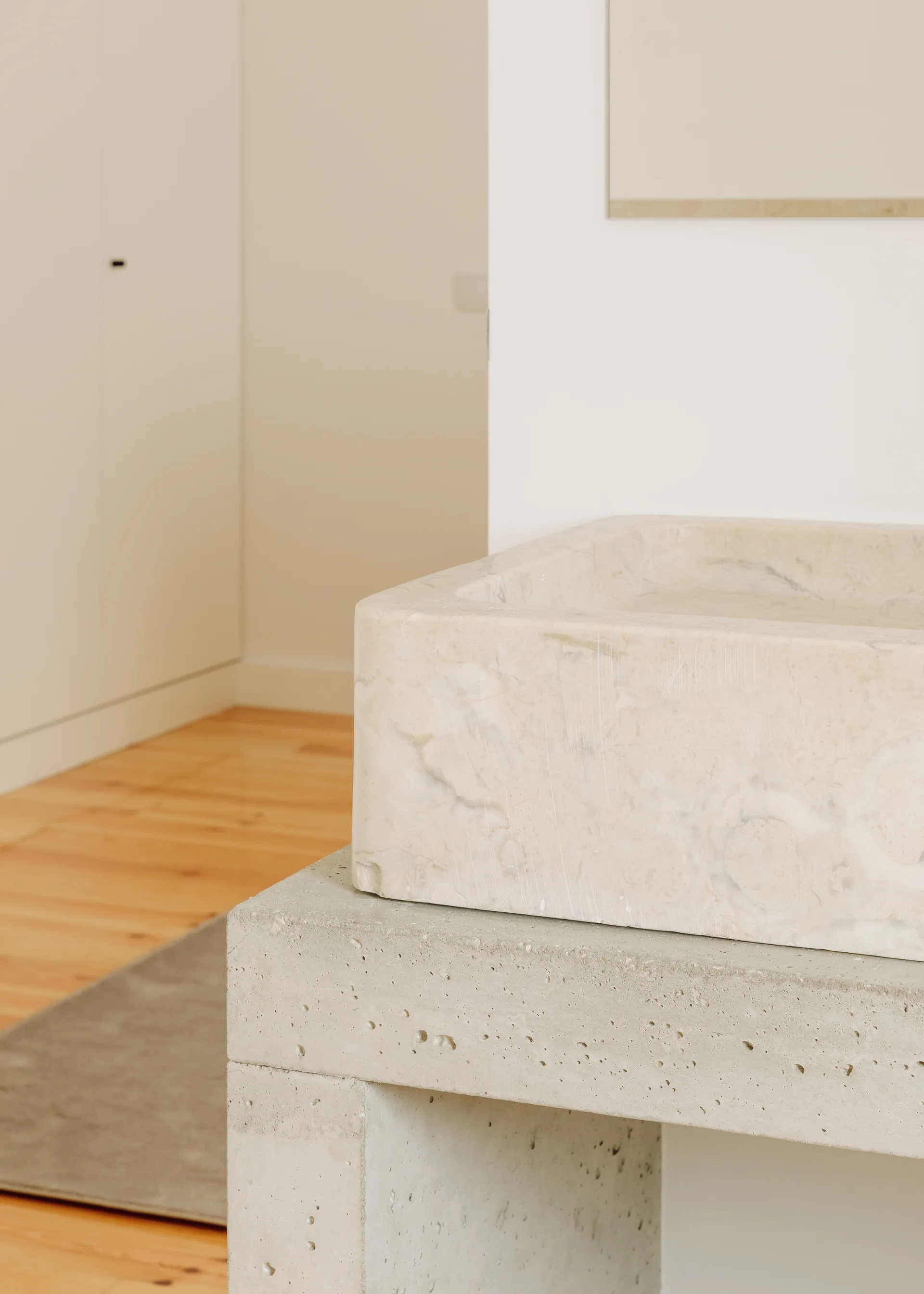 A close-up of a carved cream stone basin sitting on a grey concrete vanity base in a minimalist bathroom with wood floors.