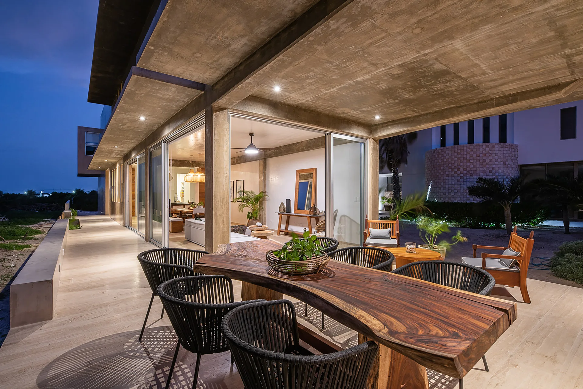 A modern outdoor concrete patio features a live-edge wood dining table with black woven chairs next to sliding glass doors opening to a warm interior.