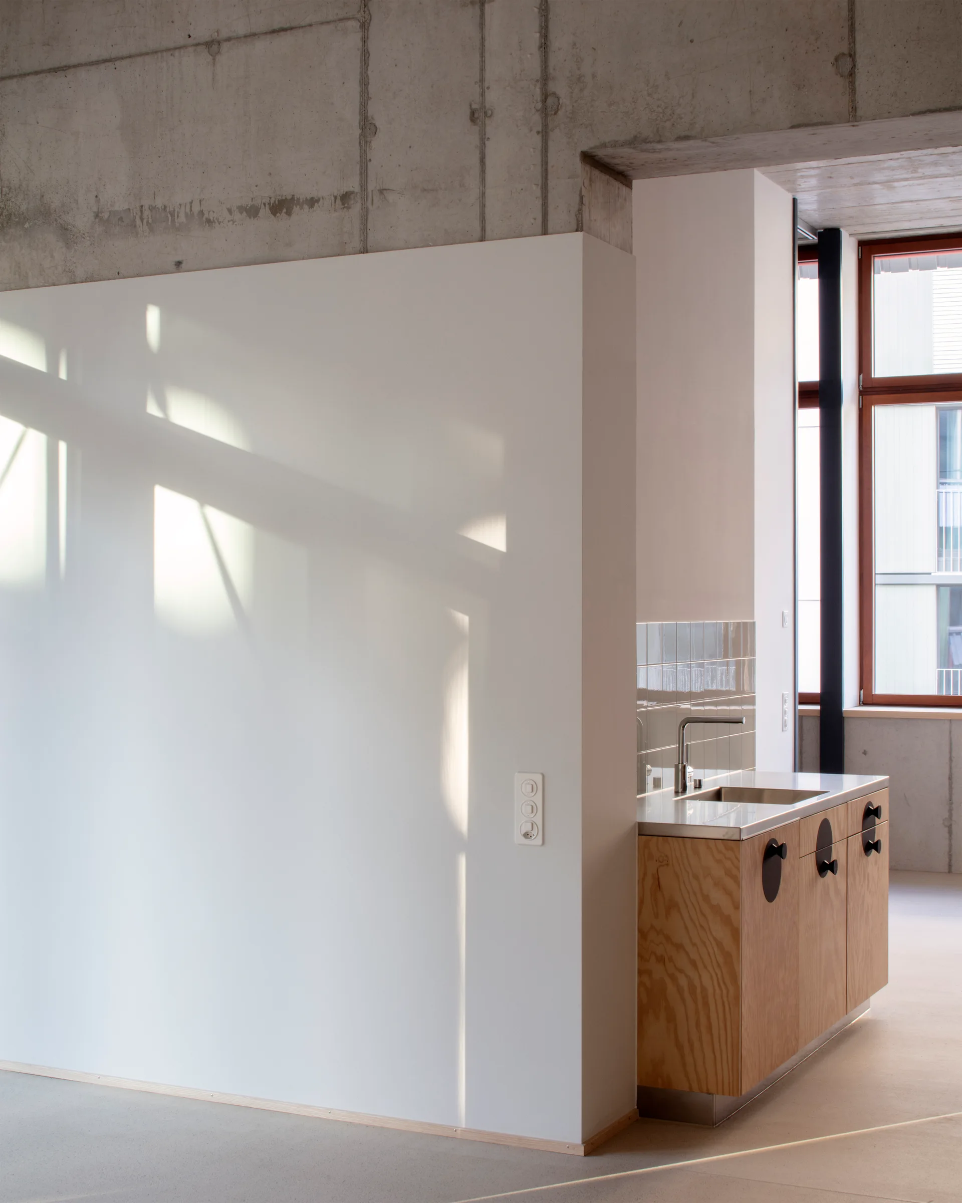 A minimalist kitchen featuring a wood grain base cabinet with a stainless steel sink, set against a white wall under an exposed concrete ceiling.