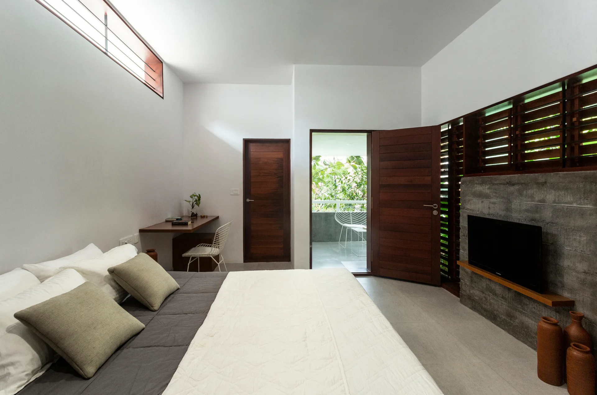 A modern bedroom featuring a bed with neutral linens, a wall-mounted wooden desk, and a television on a board-formed concrete wall.