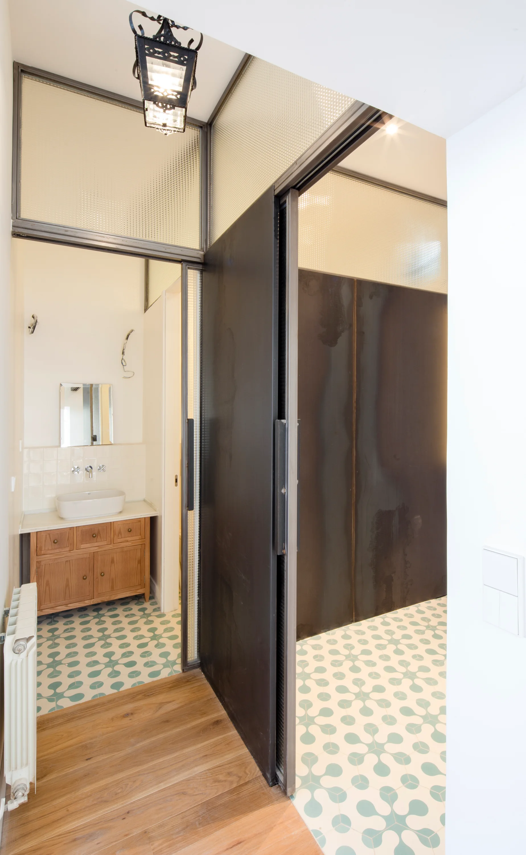 A modern bathroom view showing a green and white patterned tile floor, a wooden vanity cabinet, and a large dark metal sliding door under textured glass.