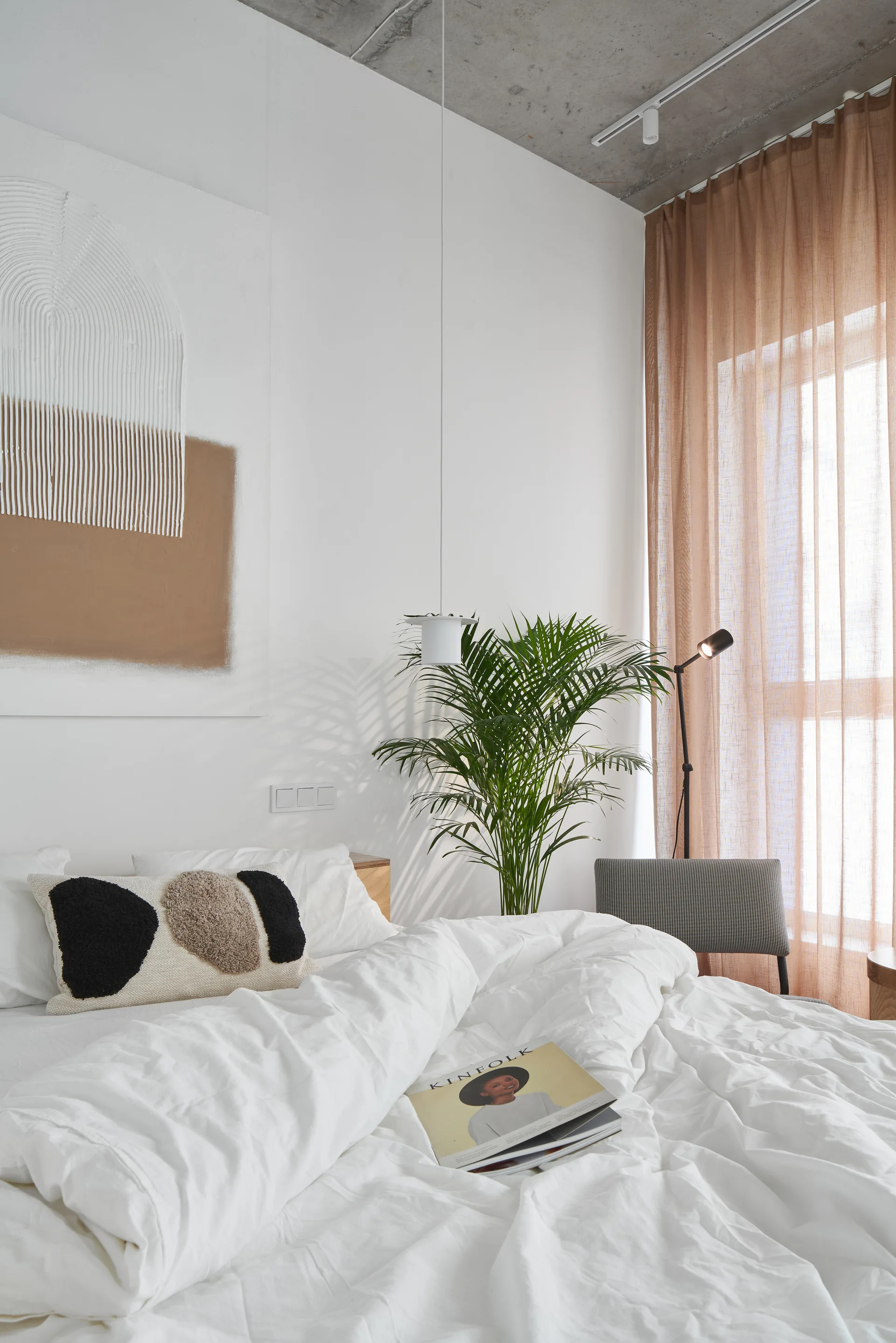 A minimalist bedroom featuring white bedding, an exposed concrete ceiling, a tall potted palm, and sheer warm-toned curtains.