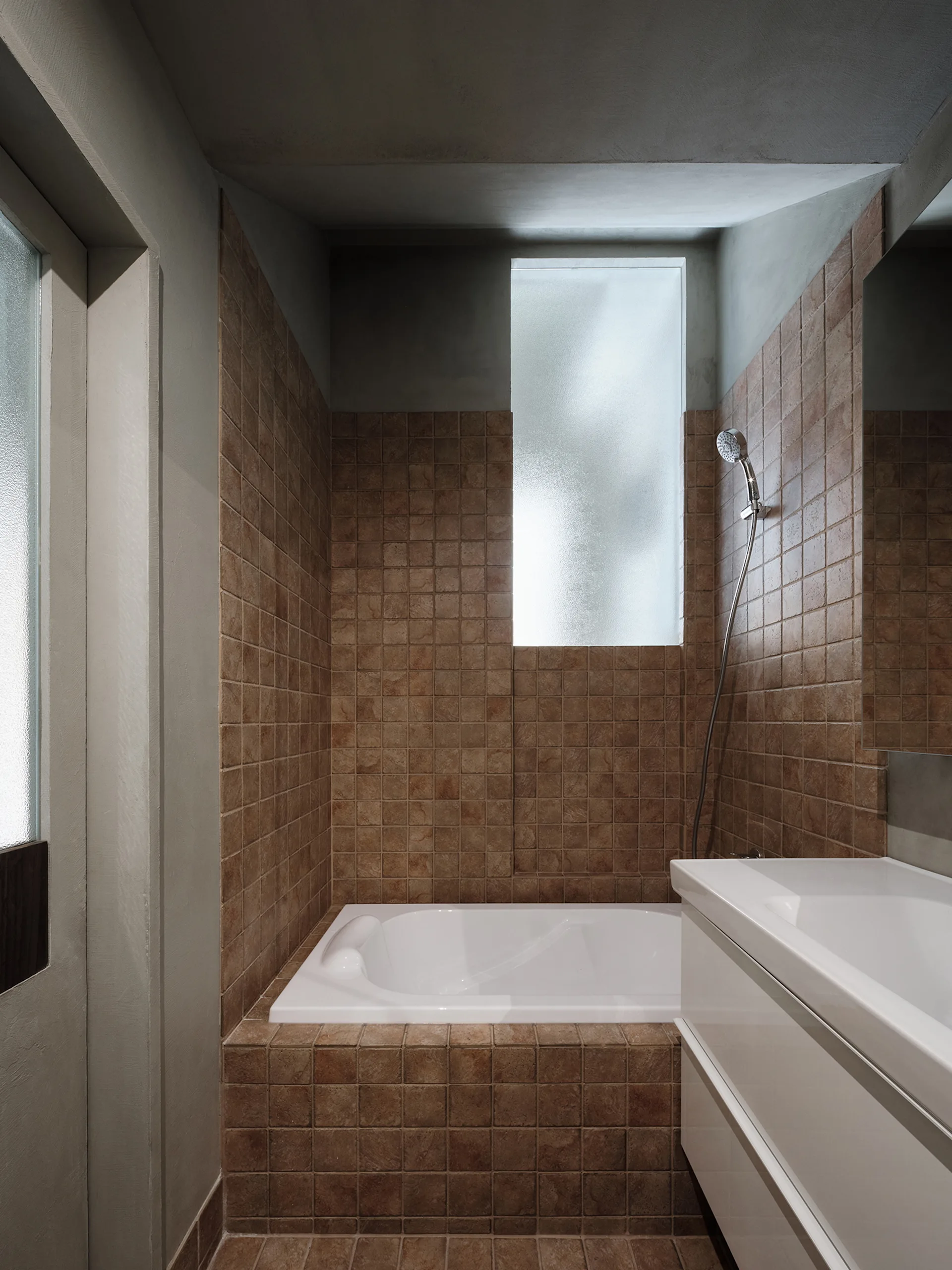A modern bathroom features a white integrated bathtub enclosed by textured brown tiles beneath a tall frosted glass window next to a white vanity.