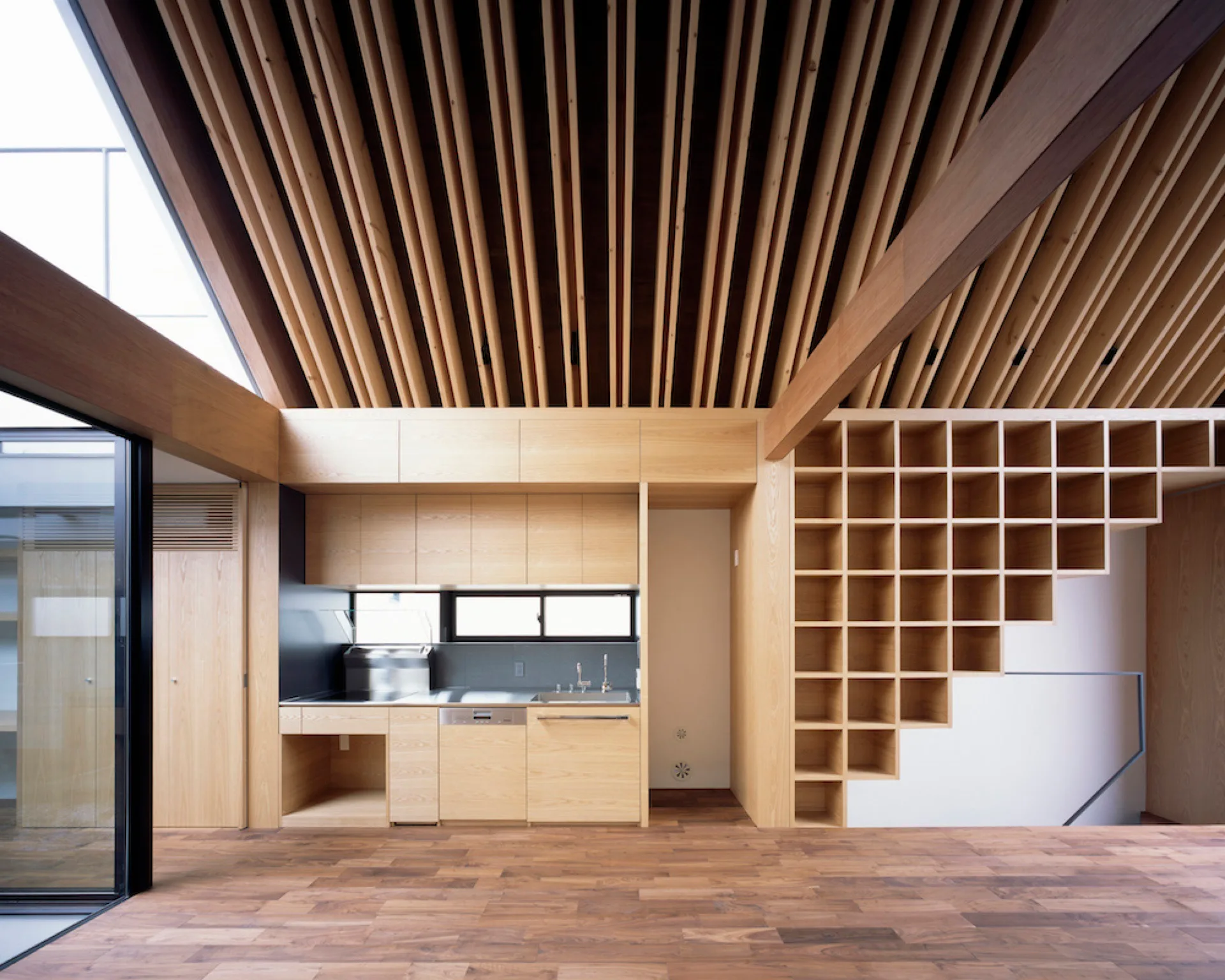 A minimalist wood kitchen featuring light timber cabinets, a matching grid-pattern storage unit, and a high ceiling made of closely spaced vertical slats.