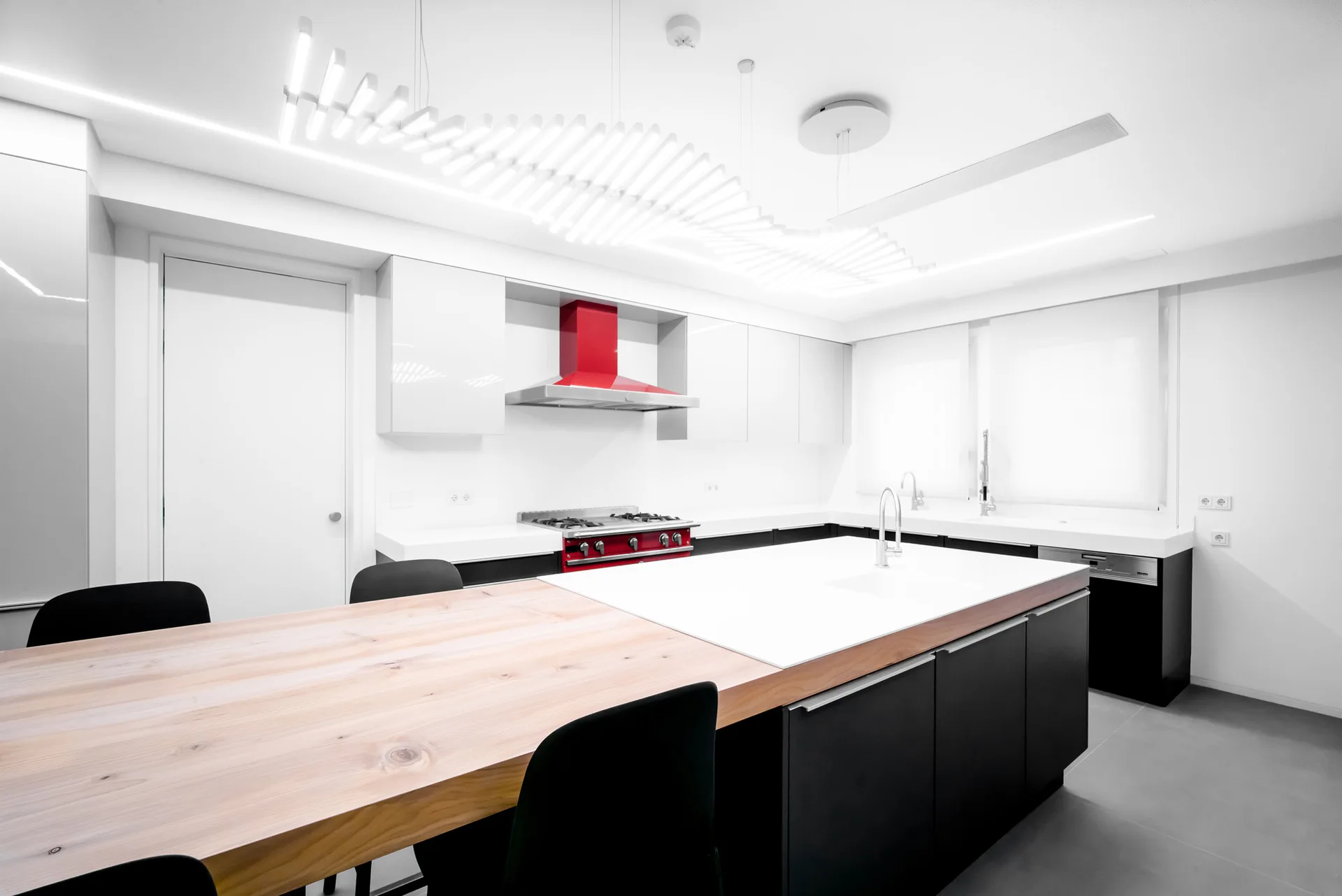 A minimalist kitchen featuring white and black cabinets, a large island with a thick wood table extension, and a red range hood.