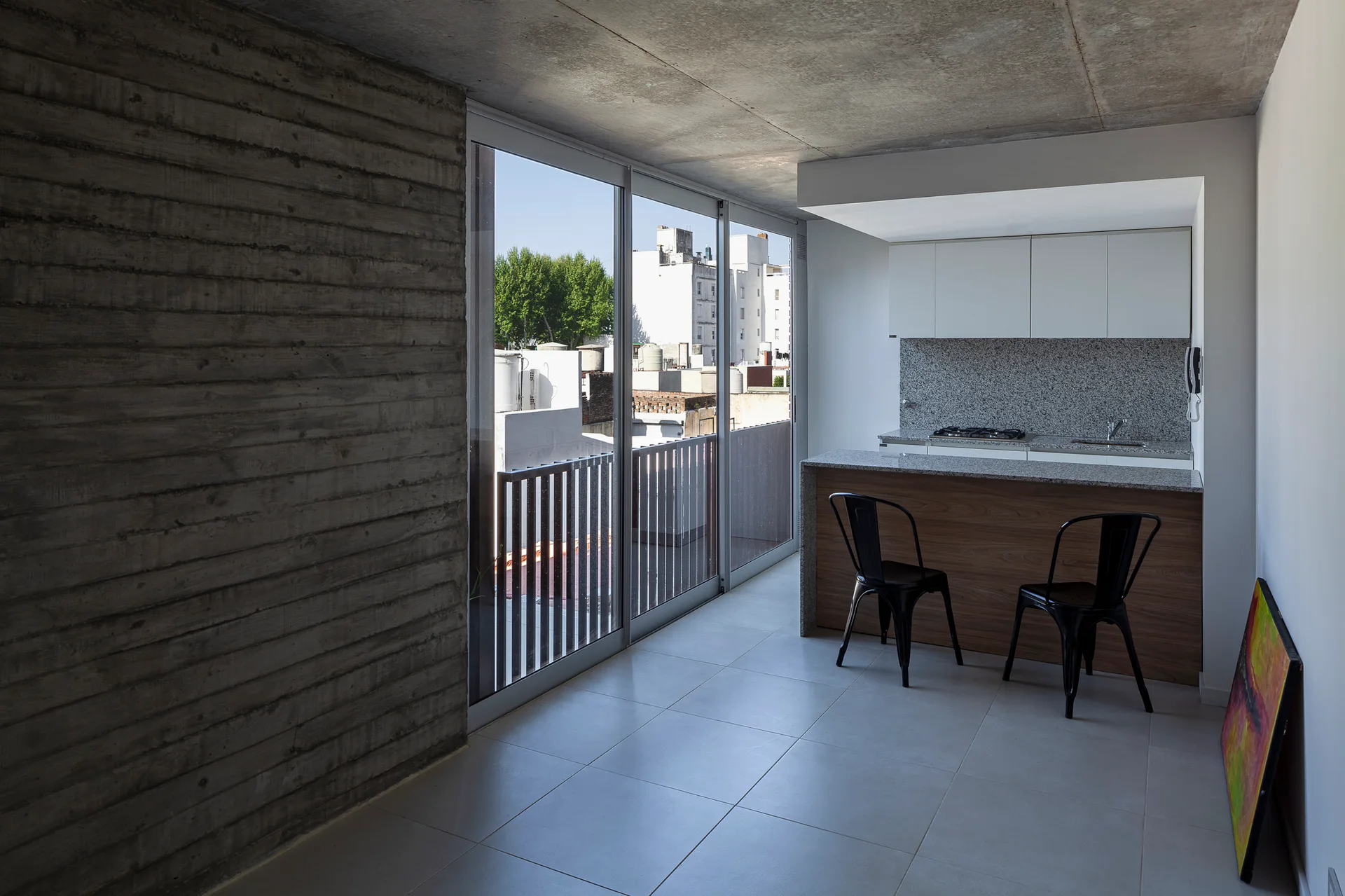 An open kitchen features a board-formed concrete wall, white cabinetry, a wood-fronted island with black stools, and sliding glass doors.