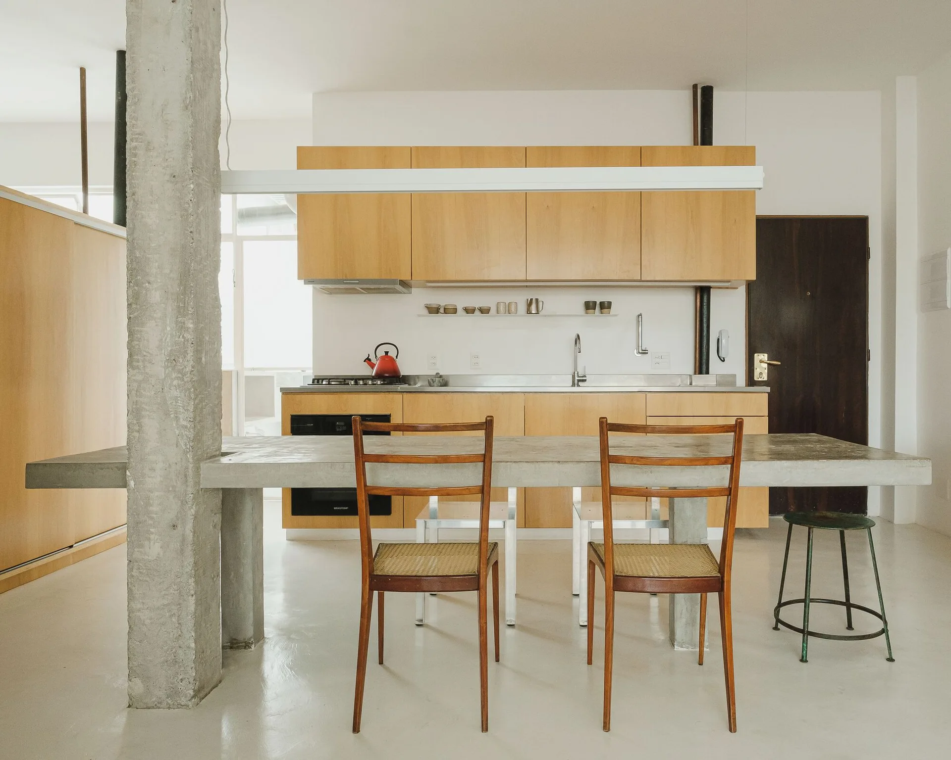 A minimalist kitchen features flat-front wood cabinets, a stainless steel counter, and a thick concrete dining table beside a raw concrete column.