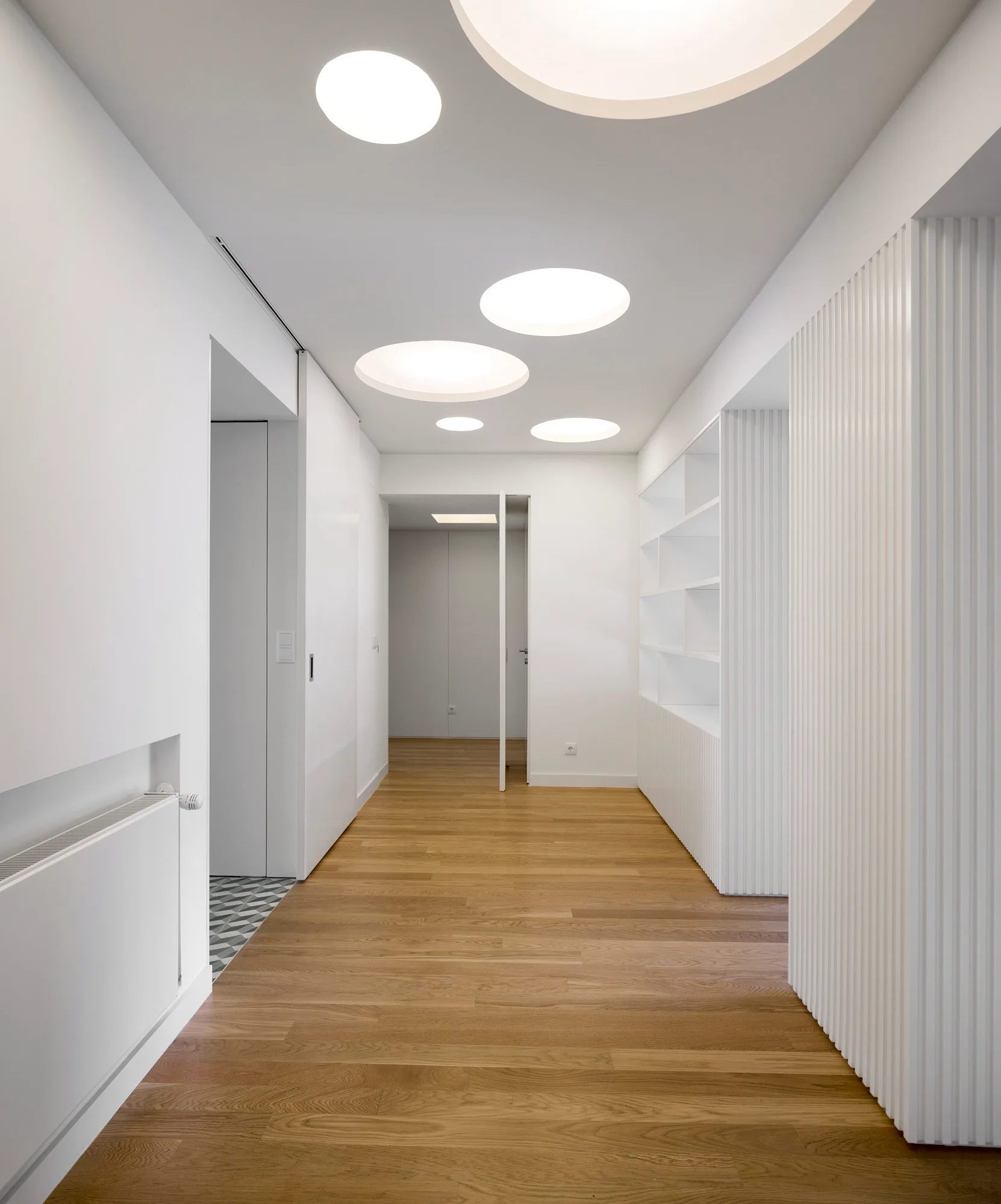 A long white hallway features light wood floors, built-in shelving, vertical fluted wall paneling, and large circular recessed ceiling lights.
