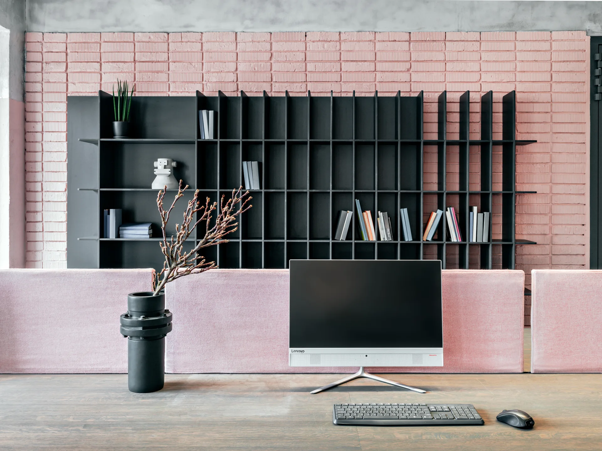 A contemporary wooden desk with an all-in-one computer stands in front of a pink partition, backed by a black grid shelving unit and painted brick wall.