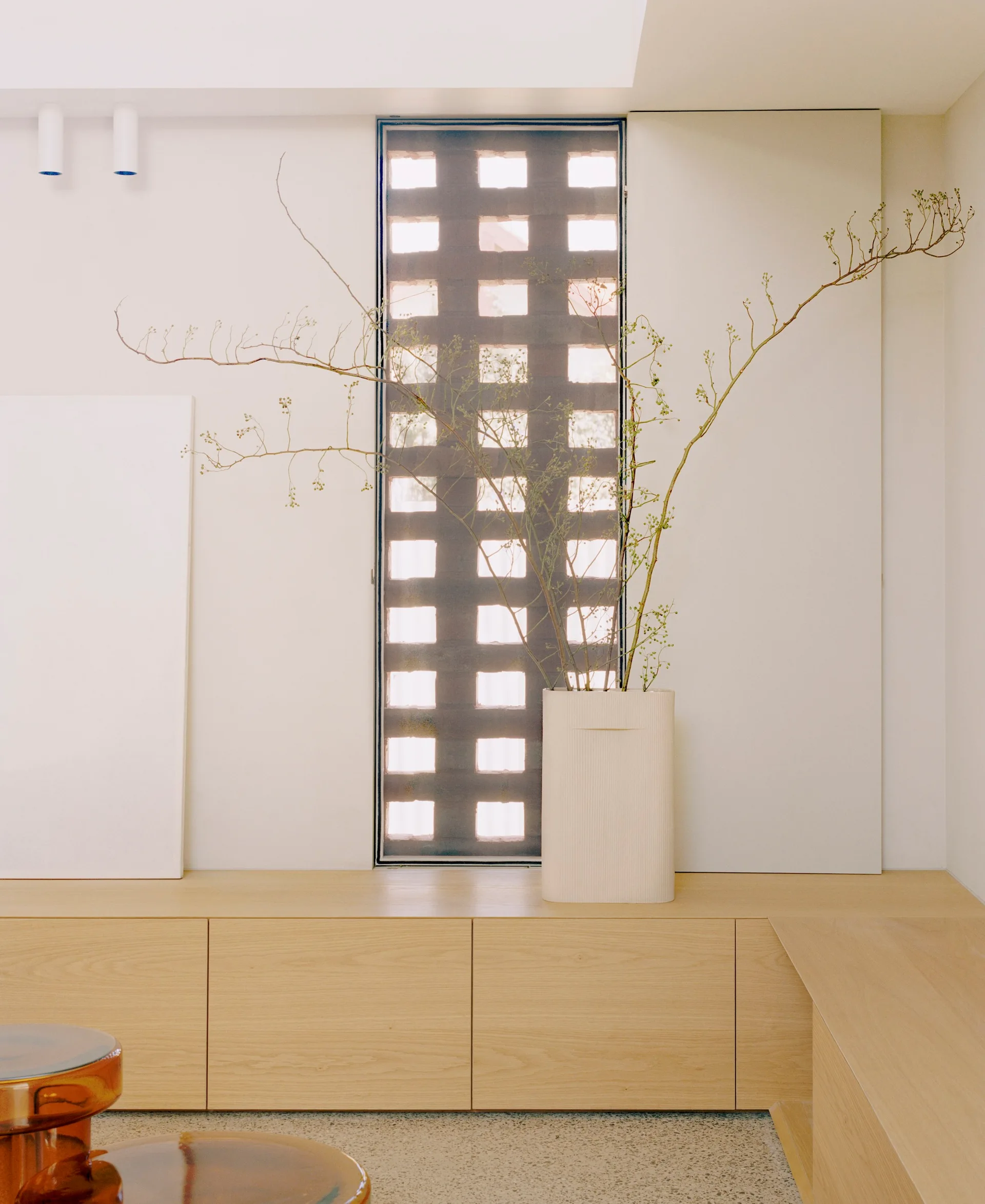 Low light wood cabinets line a white wall beneath a tall vertical grid window and a textured ceramic vase filled with wild branches.