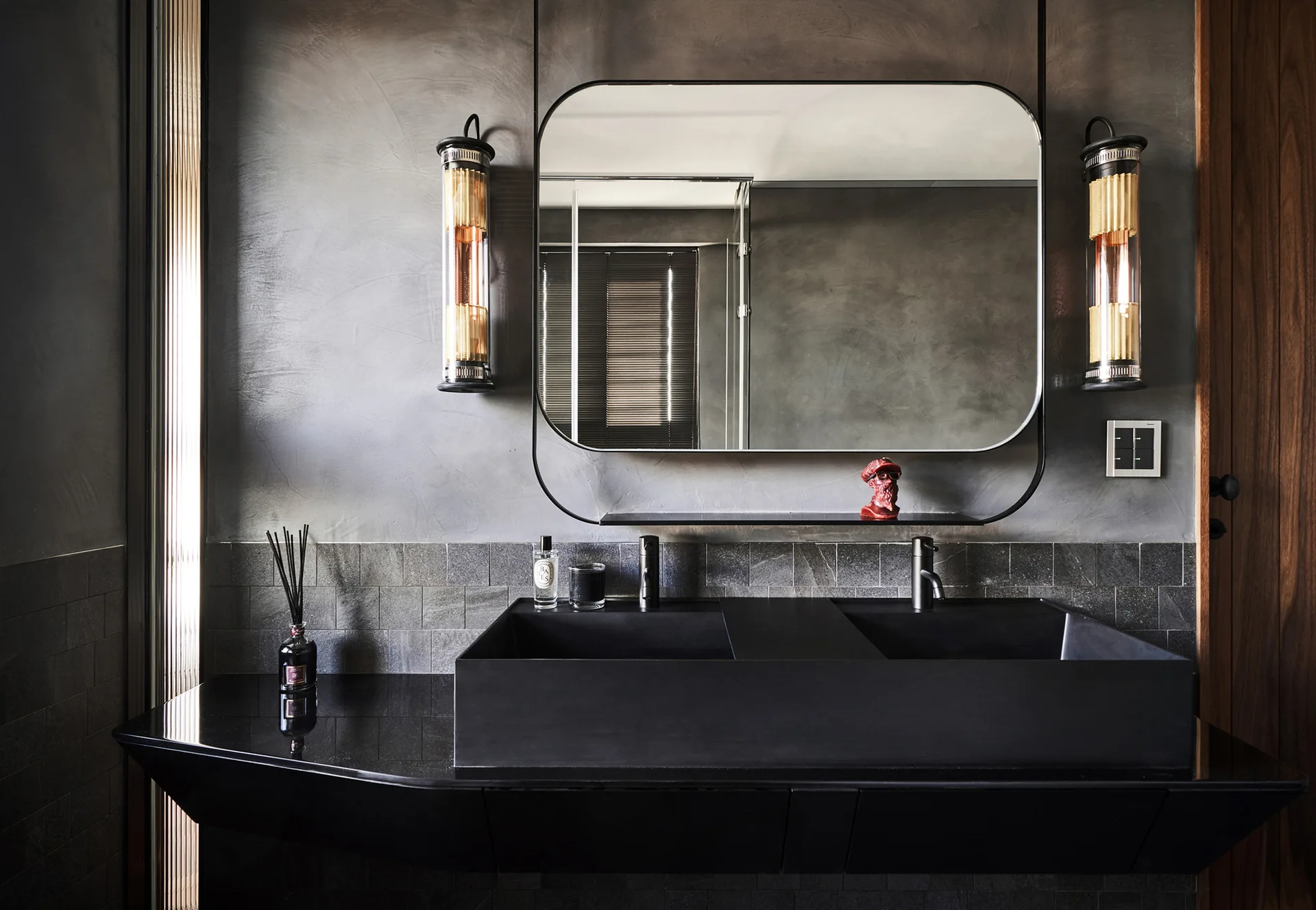A dark gray bathroom vanity features dual black sinks, a rounded rectangular mirror, and warm cylindrical wall sconces against a plaster wall.