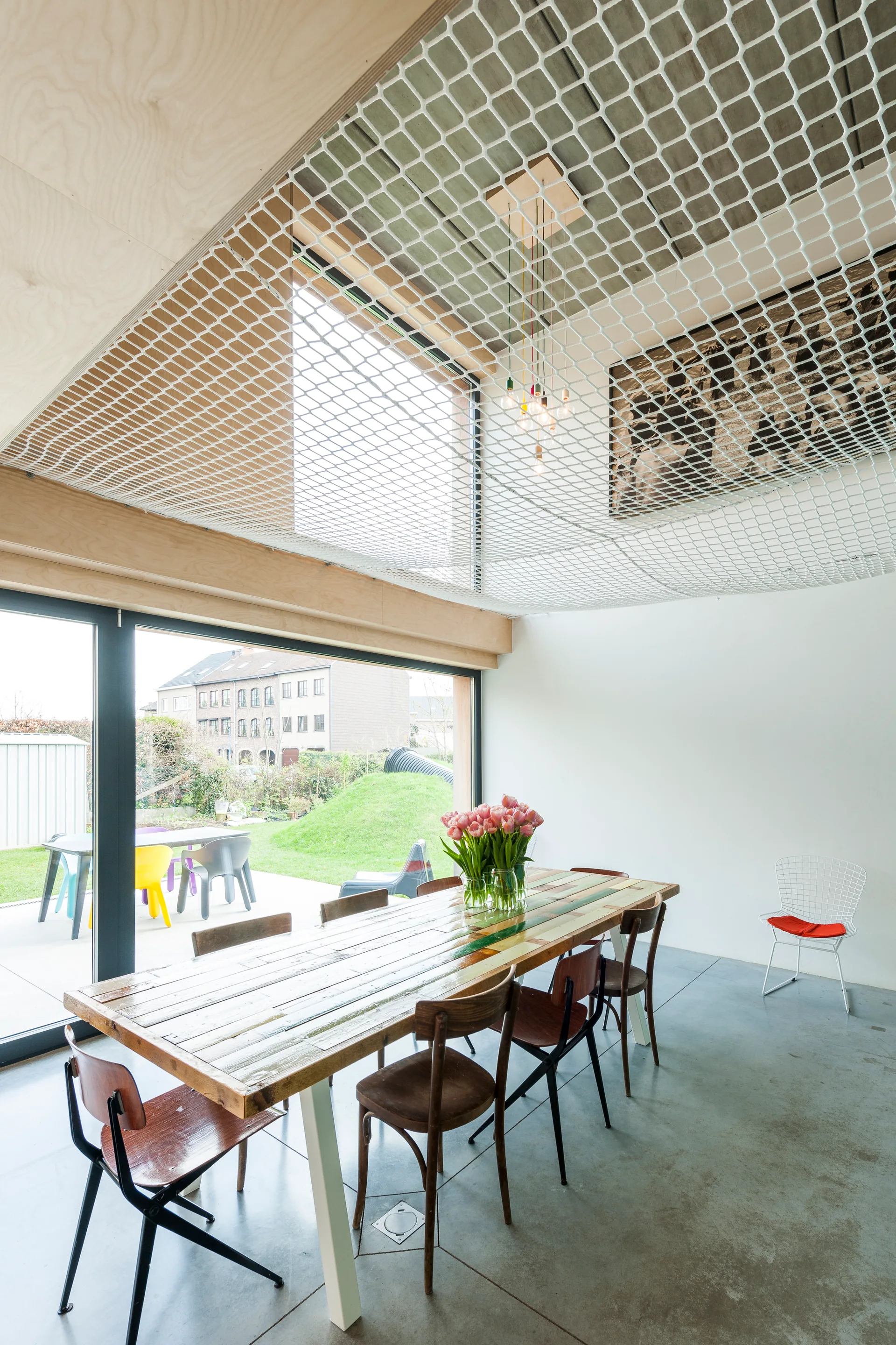 A modern dining room features a reclaimed wood dining table, mismatched vintage chairs, polished concrete floors, and a white safety net ceiling.