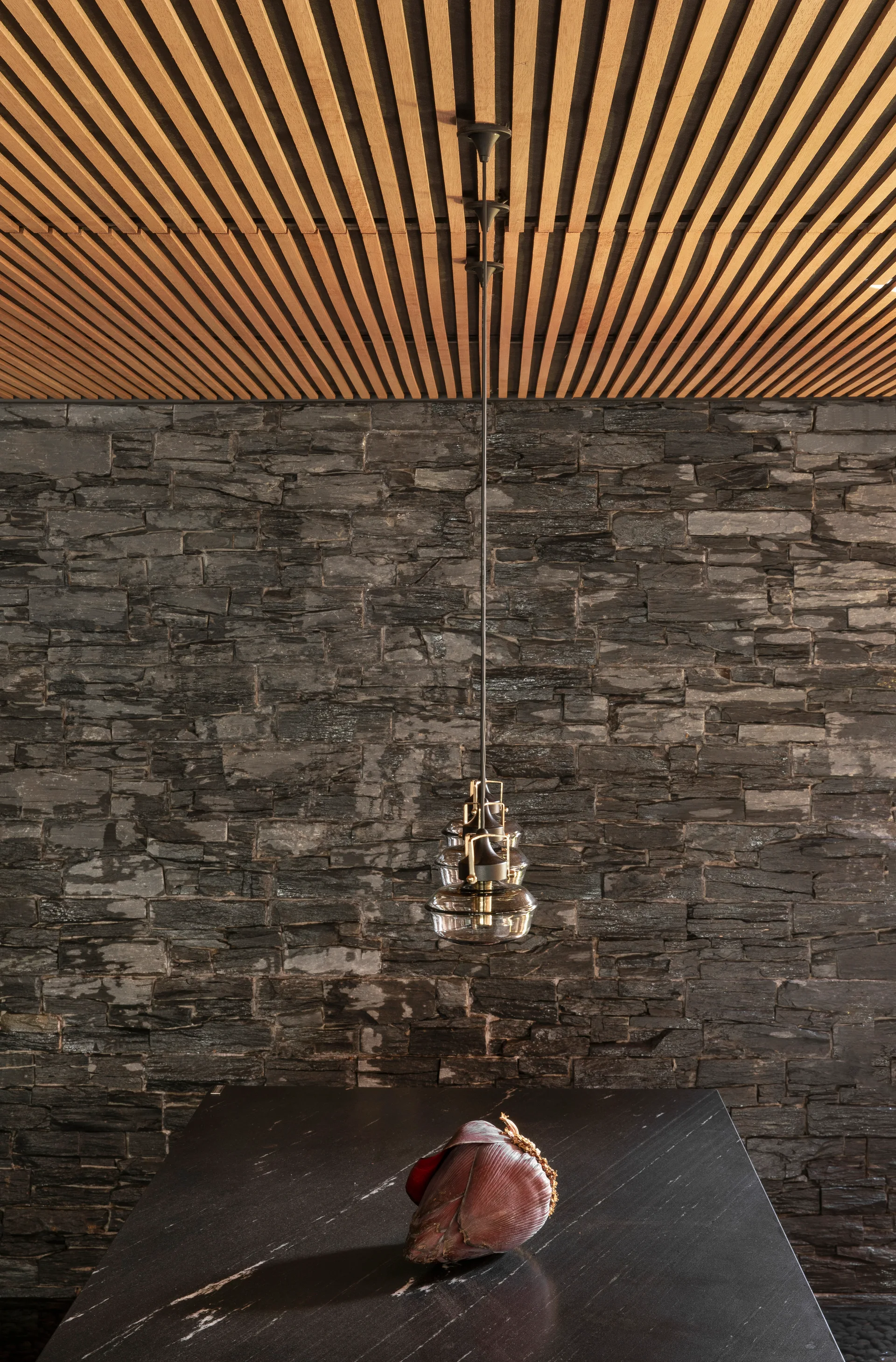 A tiered glass pendant light hangs below a linear wood slat ceiling in front of a textured dark stacked stone wall and a marble countertop.