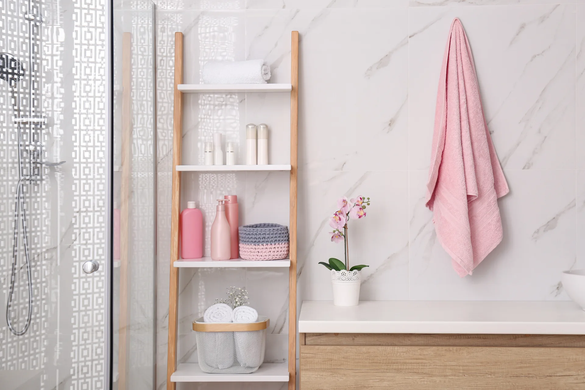 A wooden ladder shelf stocked with pink toiletries stands next to a wood-toned vanity and a hanging pink towel against a marble tile wall.