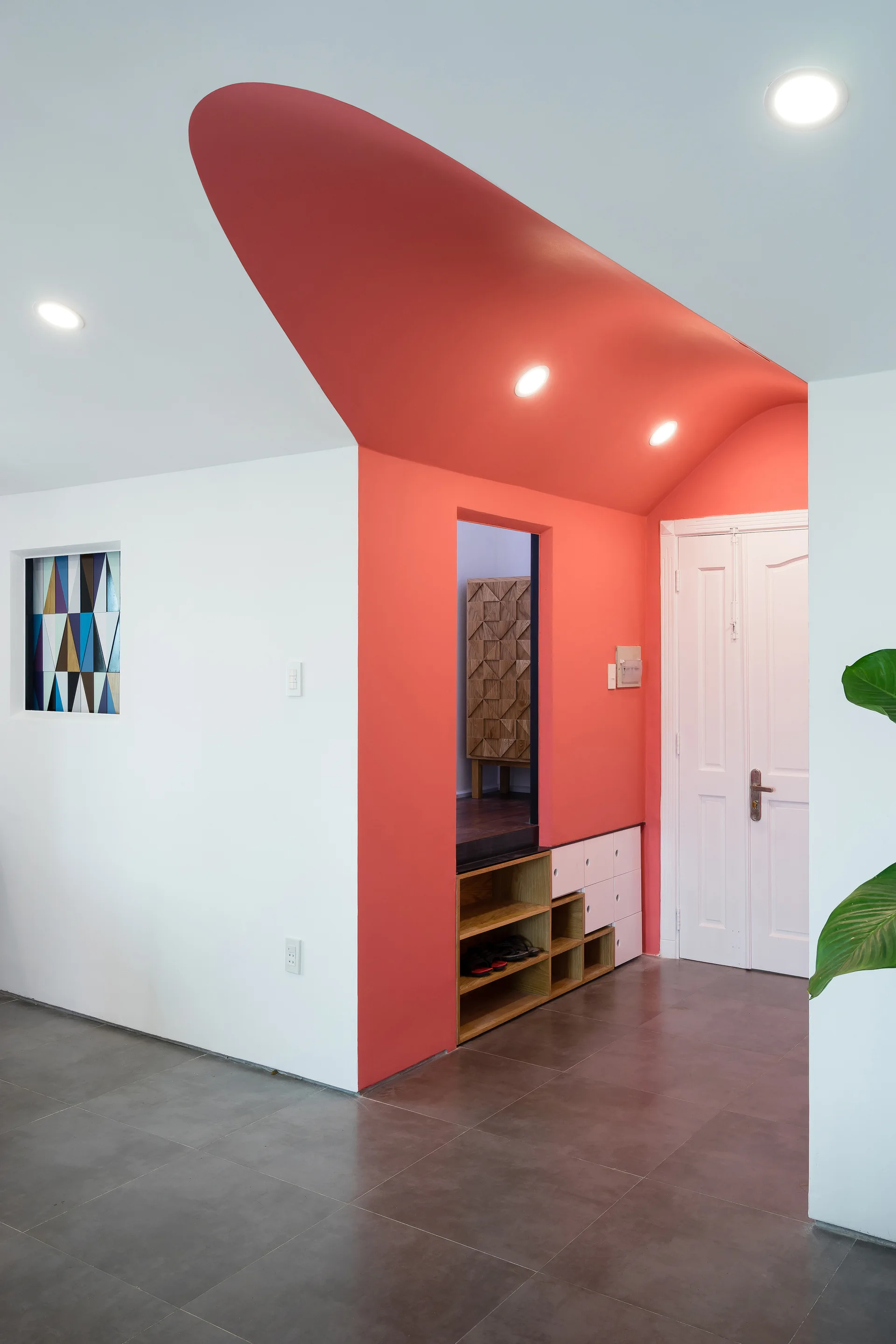 A minimalist entryway features a curved red ceiling canopy above built-in wood shoe storage next to a white door.