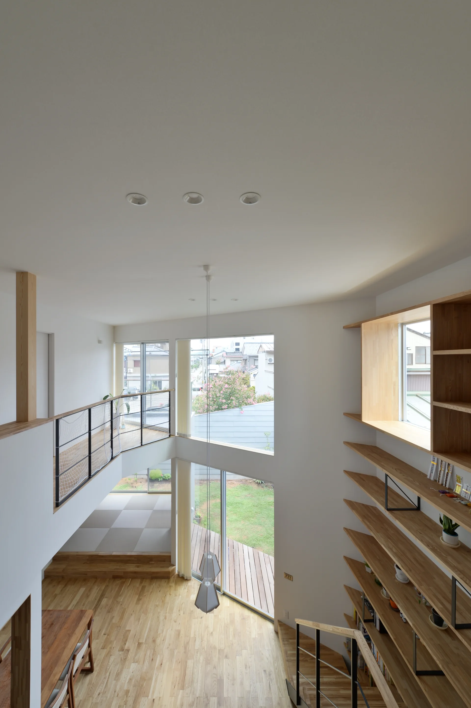 A double-height living room featuring light wood flooring, a massive built-in wooden bookshelf, and windows looking out to a backyard deck.