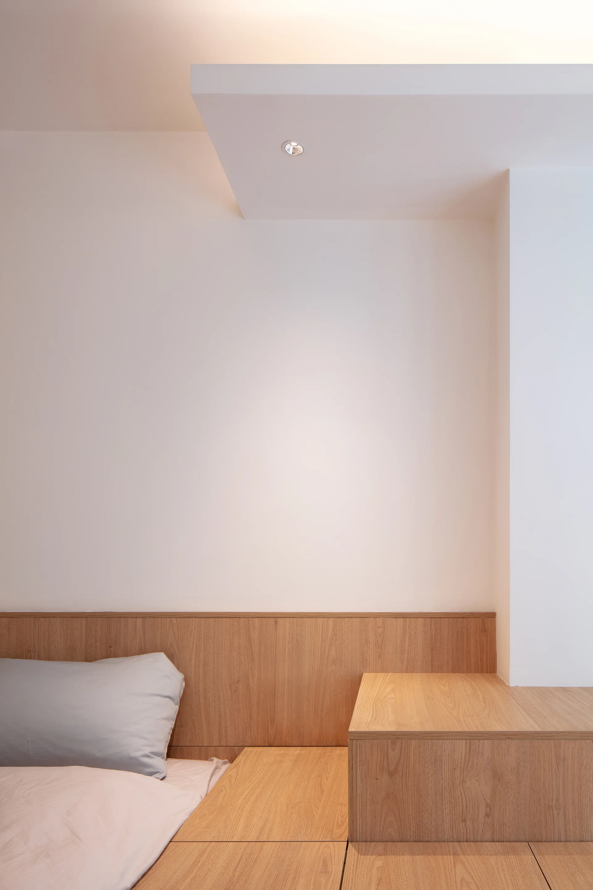 A minimalist bedroom featuring a light wood built-in platform bed and headboard panel against a plain white wall with a ceiling spotlight.