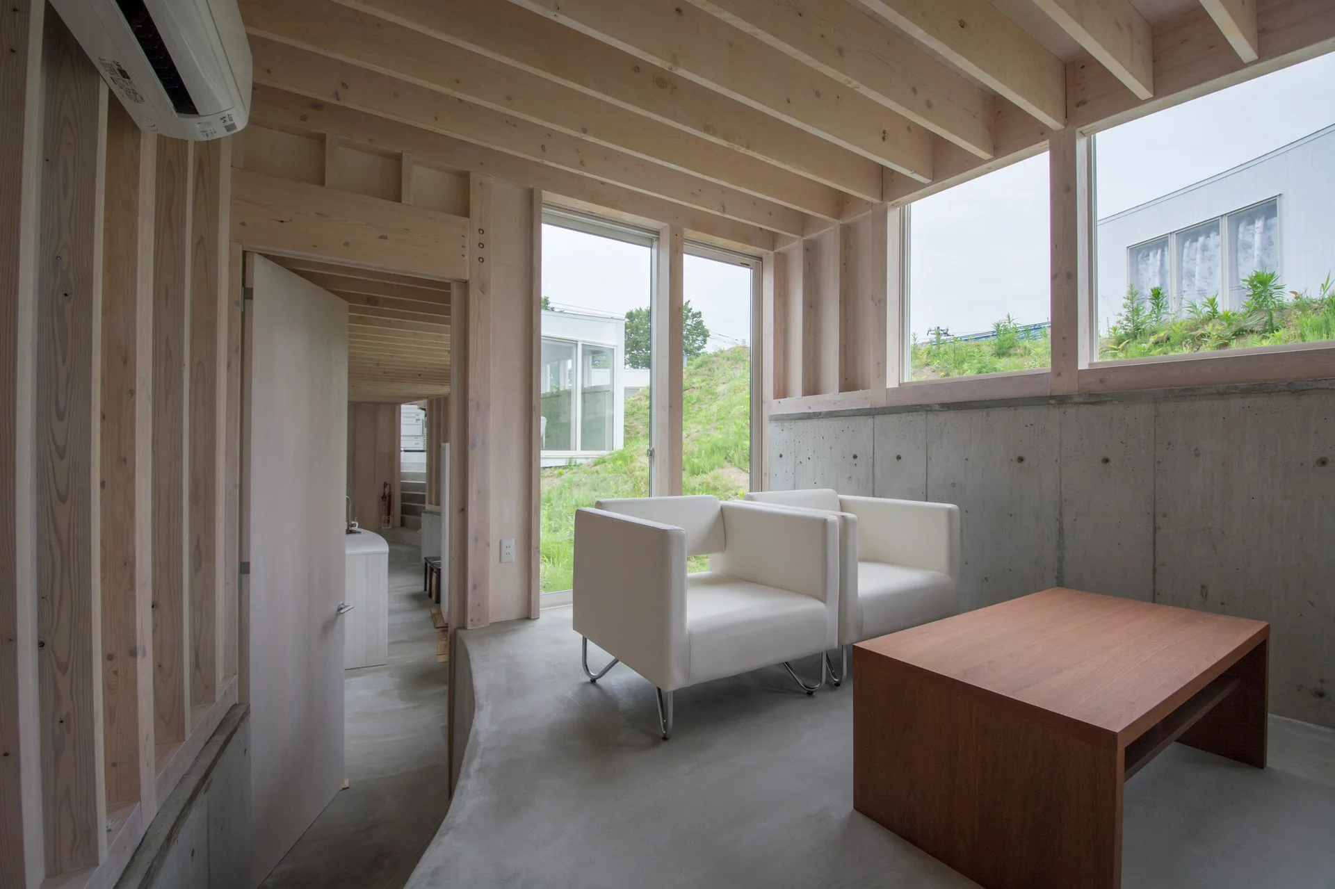 Two white armchairs and a wood coffee table sit on a concrete floor in a room with exposed timber framing and raw concrete walls.