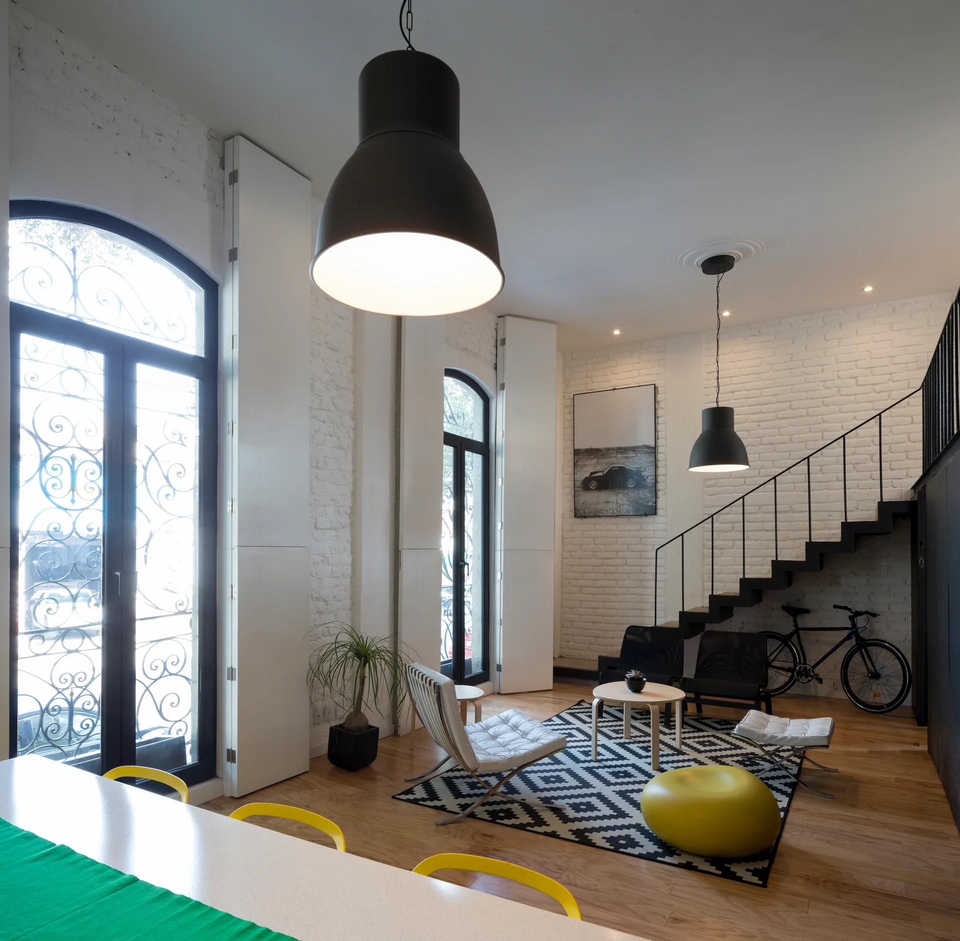 An industrial loft living room with white painted brick walls, a black metal staircase, two large black pendant lights, and a geometric area rug.