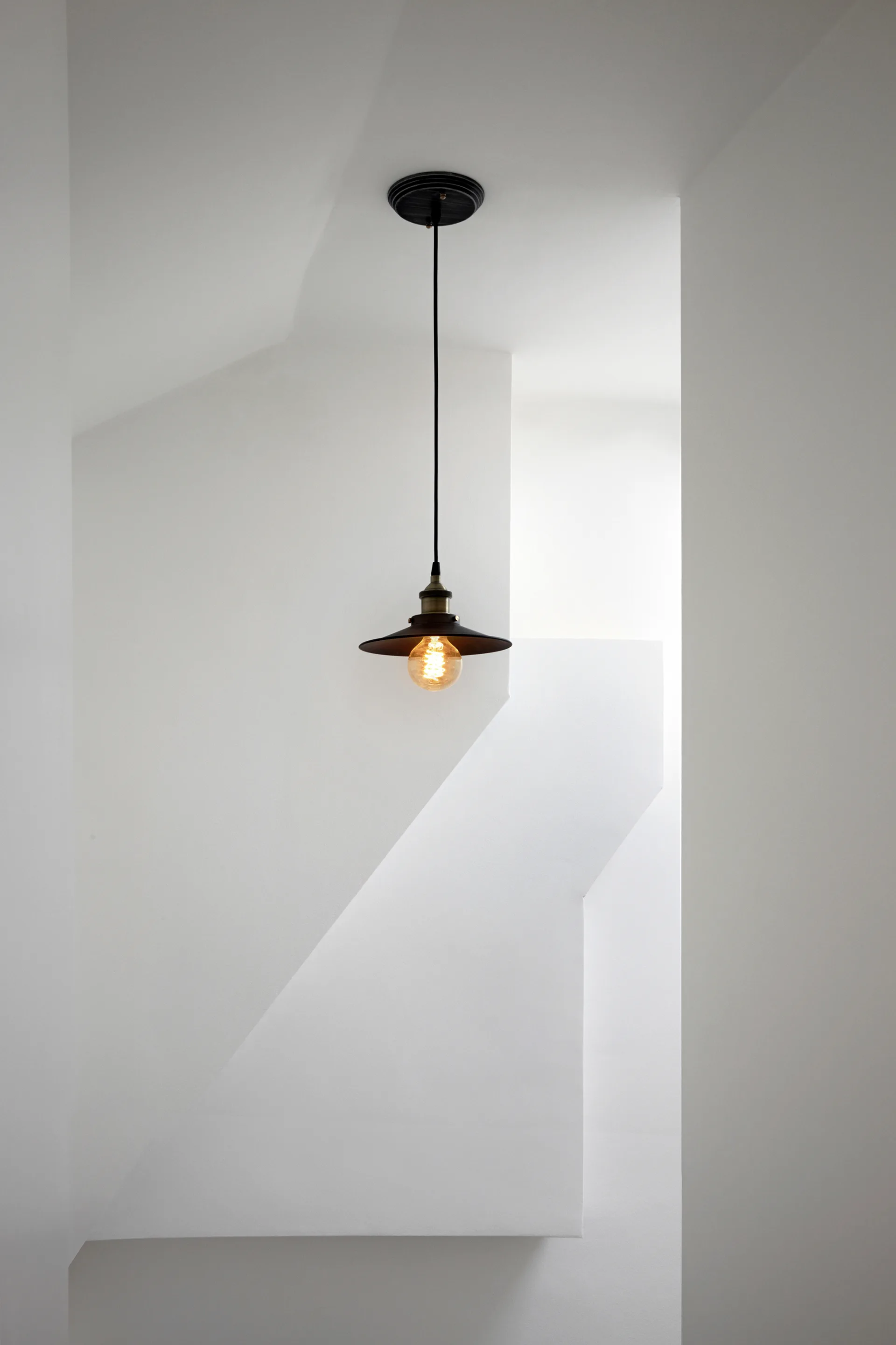 A black metal pendant light with a glowing filament bulb hangs against sculptural white staircase walls.