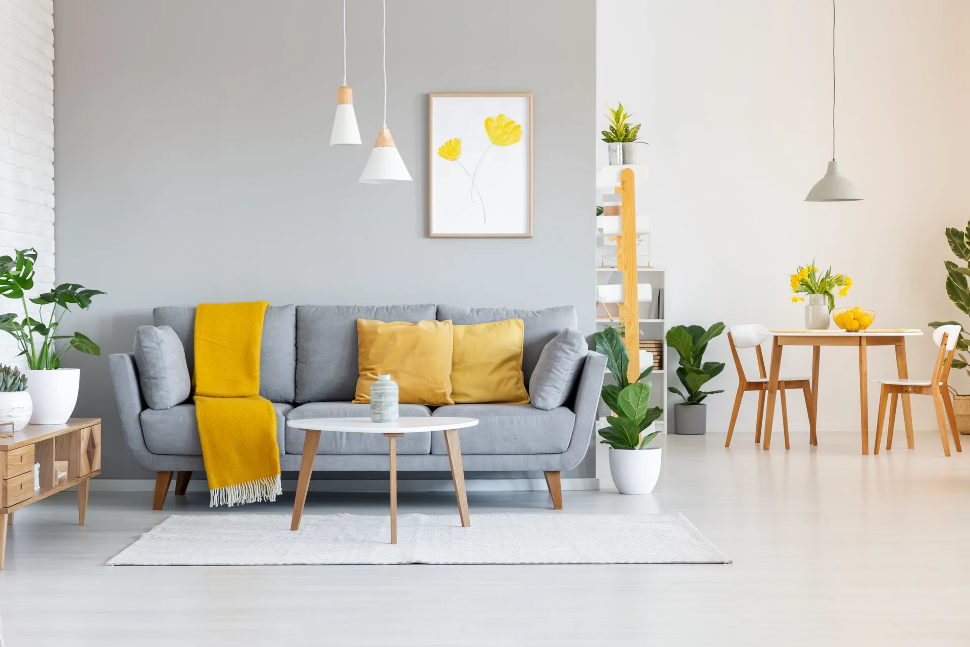 A light gray sofa with bright yellow cushions sits on a white rug in an open-plan room with a dining set in the background.