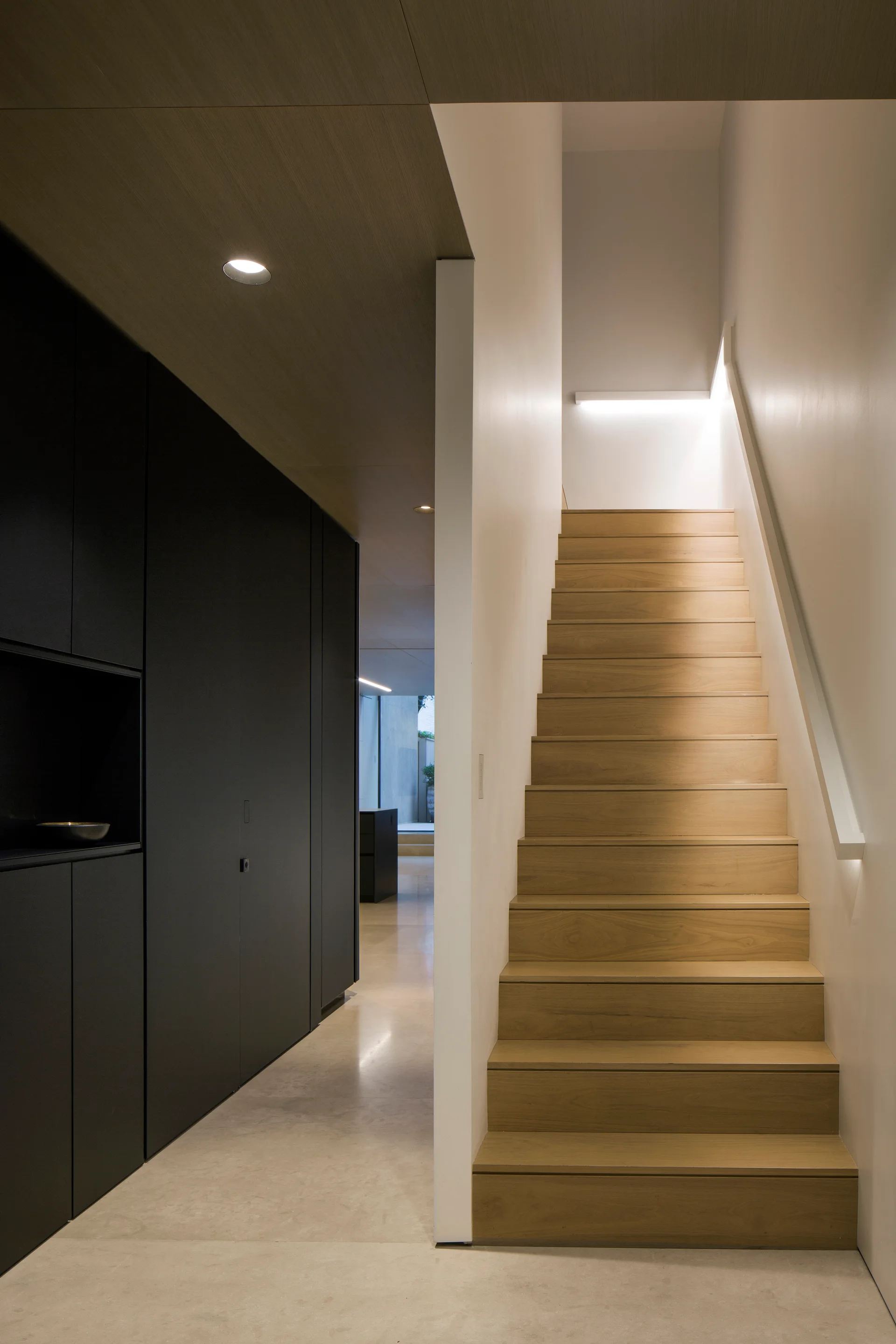 A minimalist modern hallway features a light wood staircase alongside full-height matte black cabinetry and a polished concrete floor.