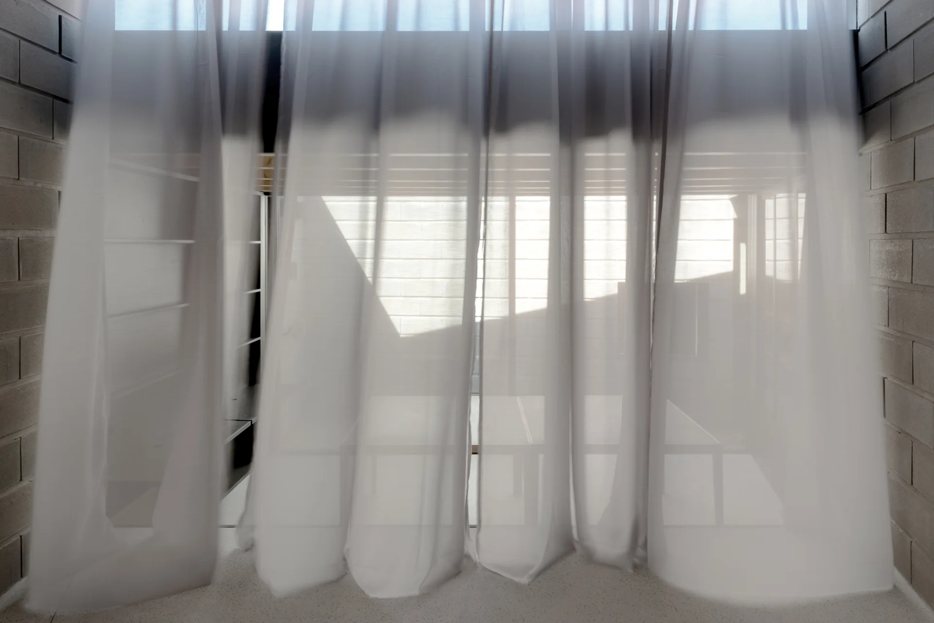 White sheer curtains hang between two exposed gray cinder block walls, filtering soft light into a minimalist room.