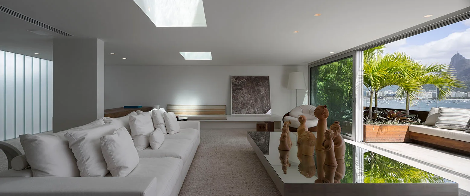 A minimalist white living room features a long low sofa, a mirrored coffee table with clay sculptures, and sliding glass doors opening to a tropical balcony.