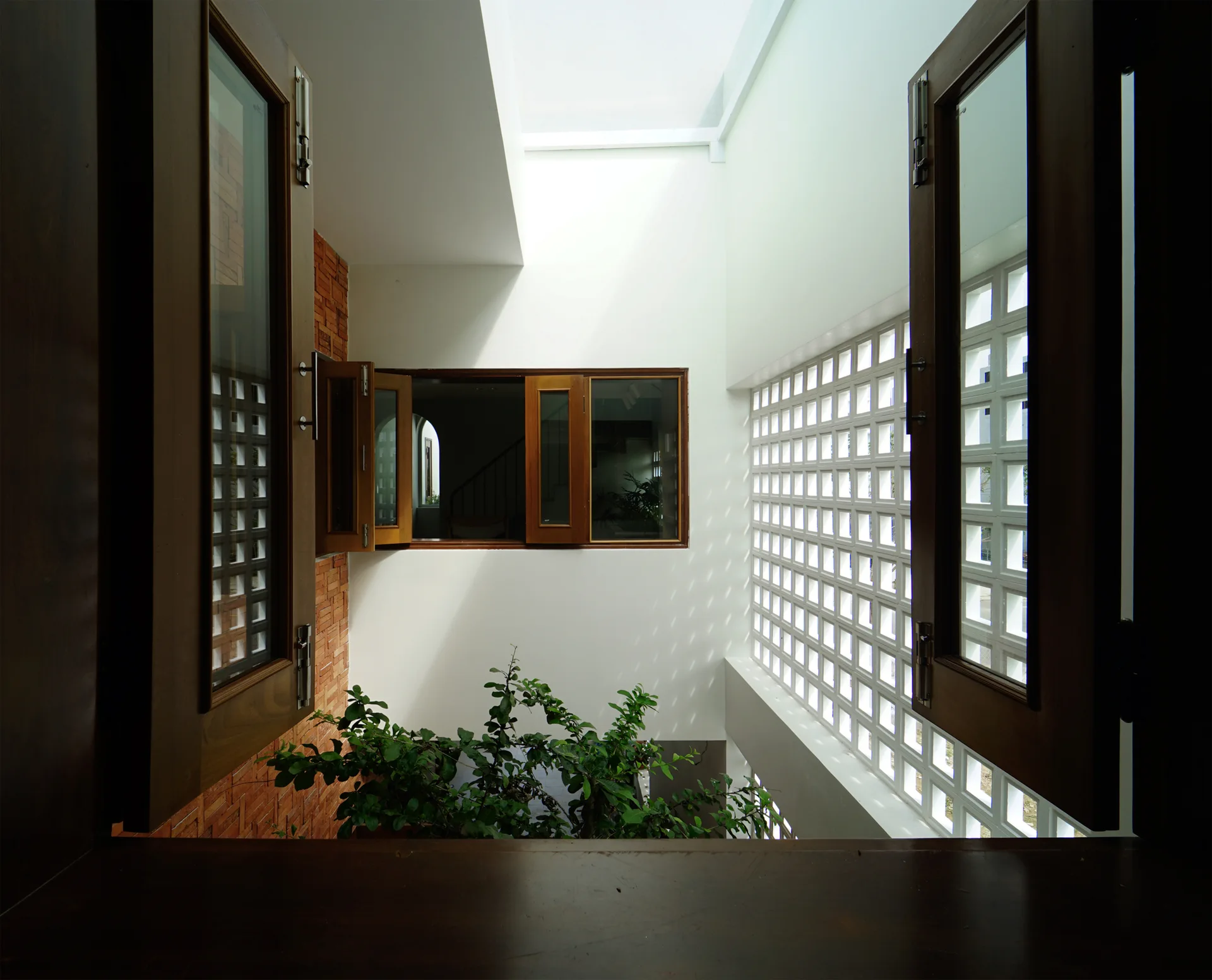 An interior view through dark wood casement windows looking into a bright internal courtyard with a white breezeblock wall and red brickwork.
