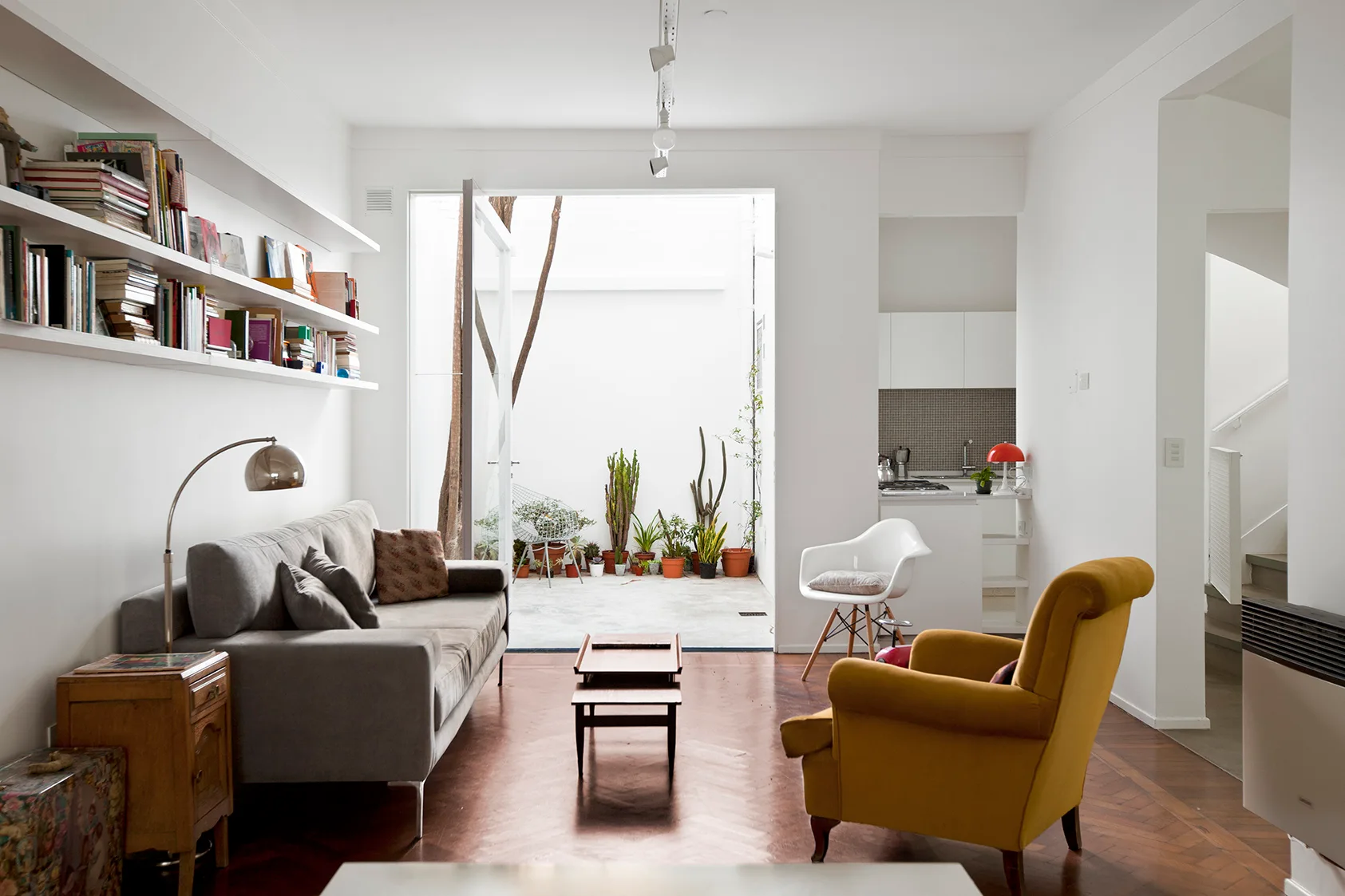 A living room featuring a gray sofa below built-in white bookshelves, a yellow armchair, herringbone wood floors, and an open doorway to a courtyard.