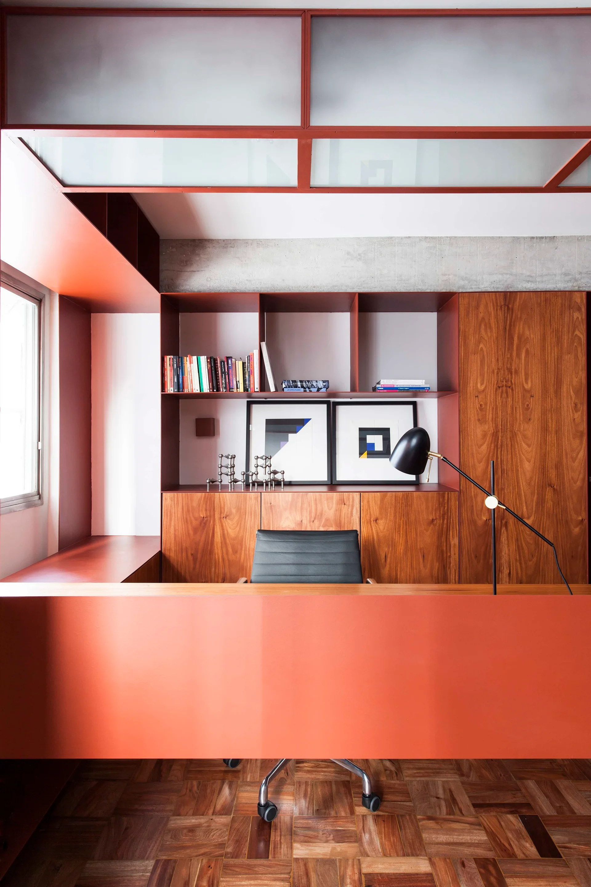 A modern home office features a terracotta orange built-in desk and shelving unit, rich wood grain cabinets, parquet flooring, and a black chair.
