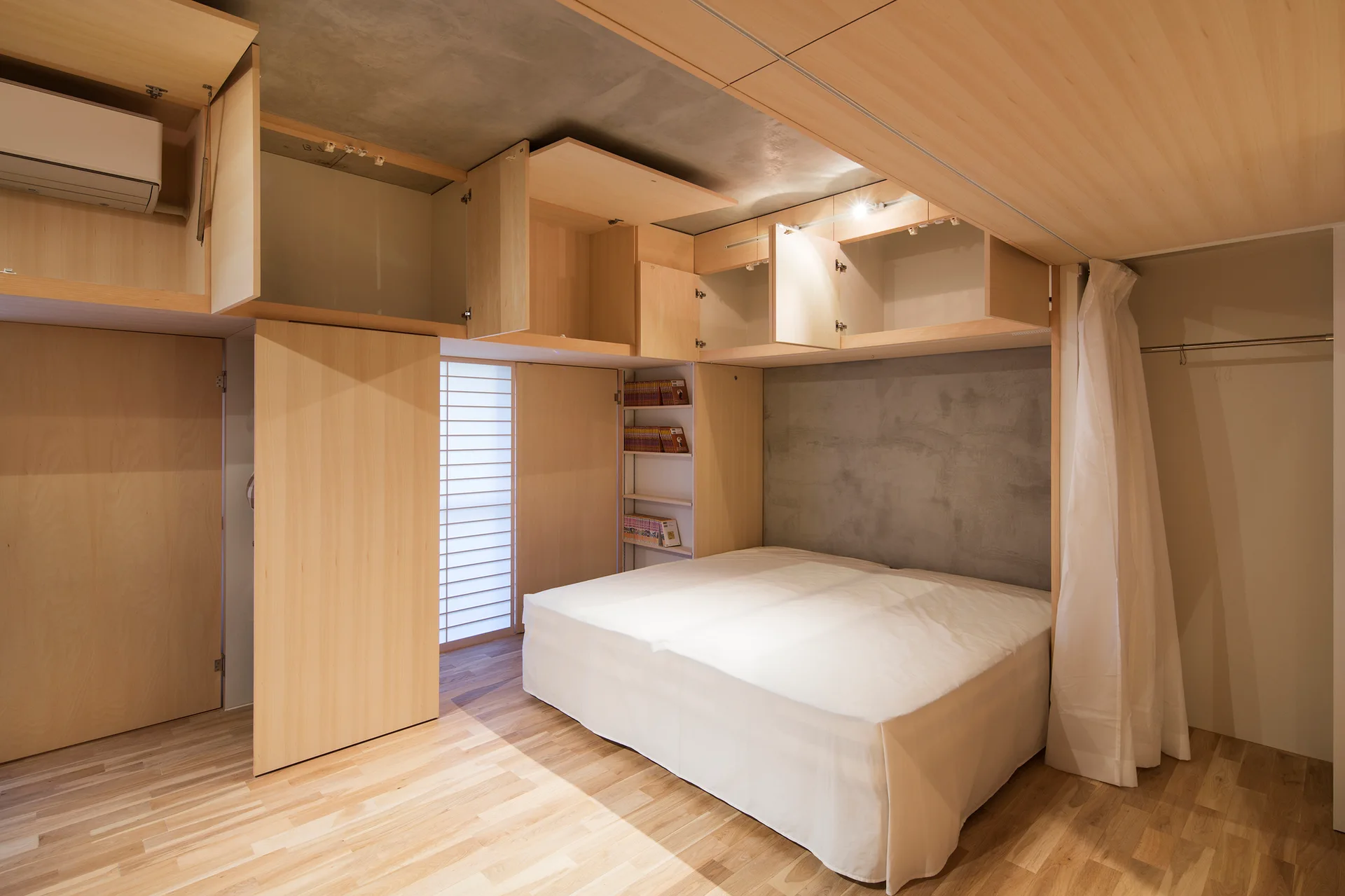 A minimalist bedroom featuring a bed set against a raw concrete wall, surrounded by light wood built-in cabinets and wood flooring.