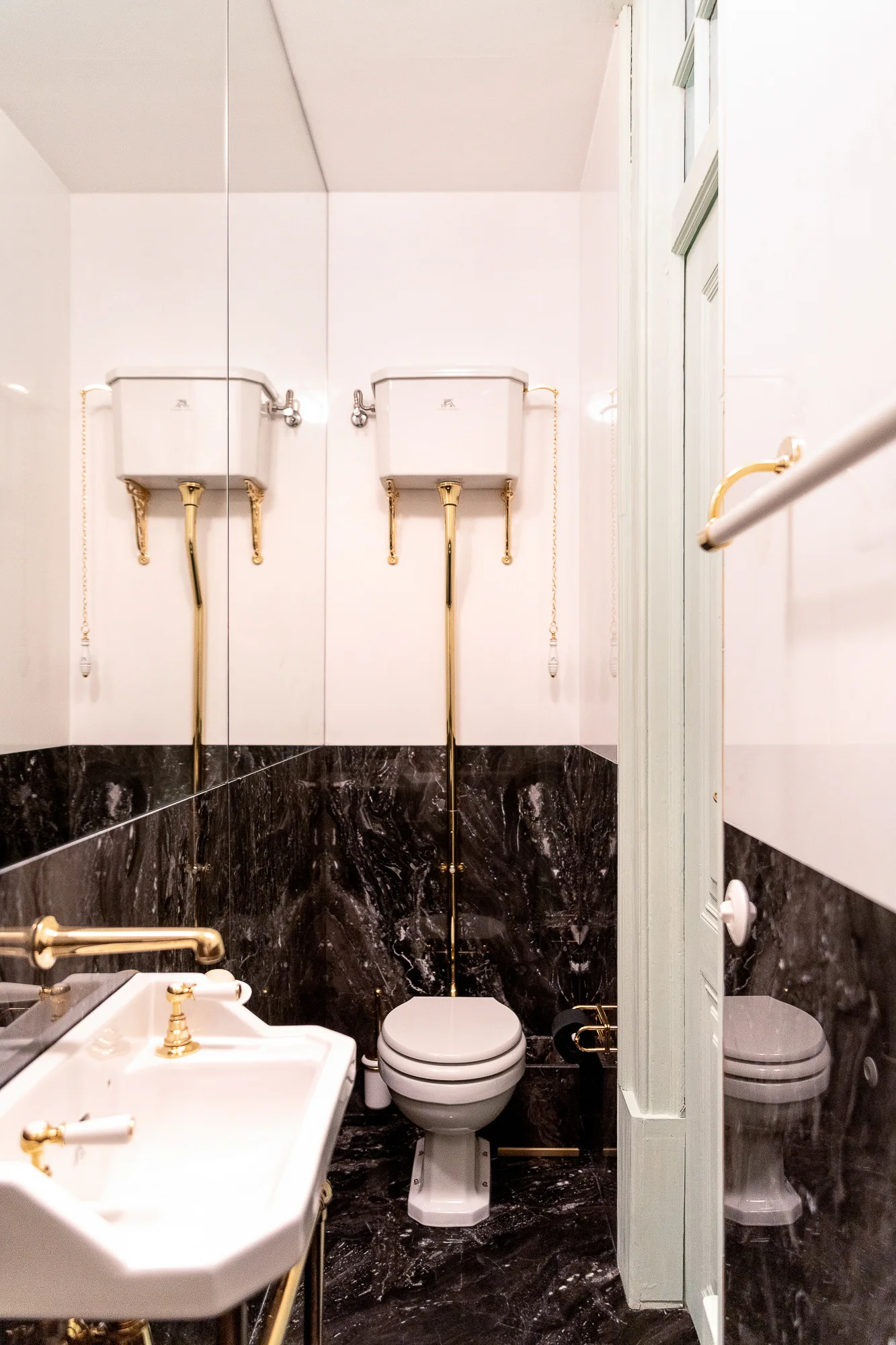 A small powder room features a white high-cistern toilet with gold pipes, a matching ceramic sink, and dark black marble wainscoting.