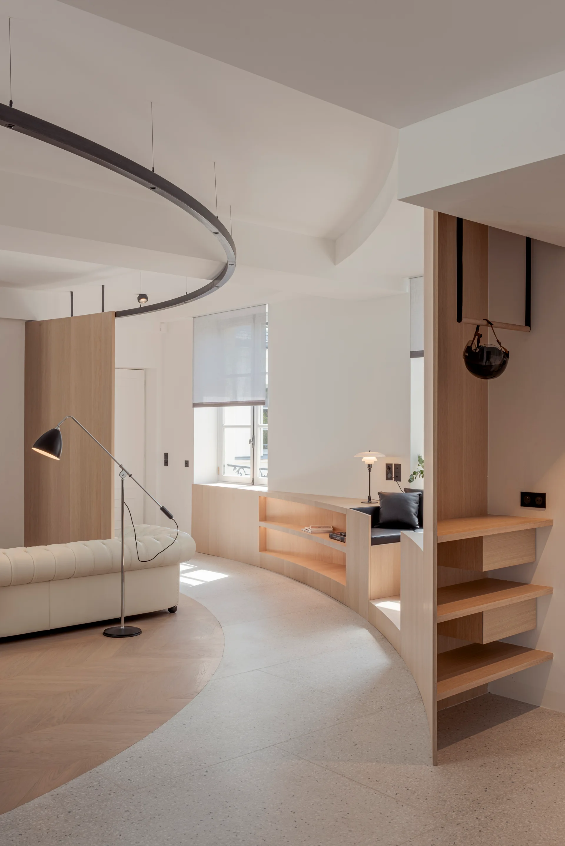 A minimalist living room showing a curved wooden built-in bookcase, a cream tufted sofa, and a circular overhead ceiling track.