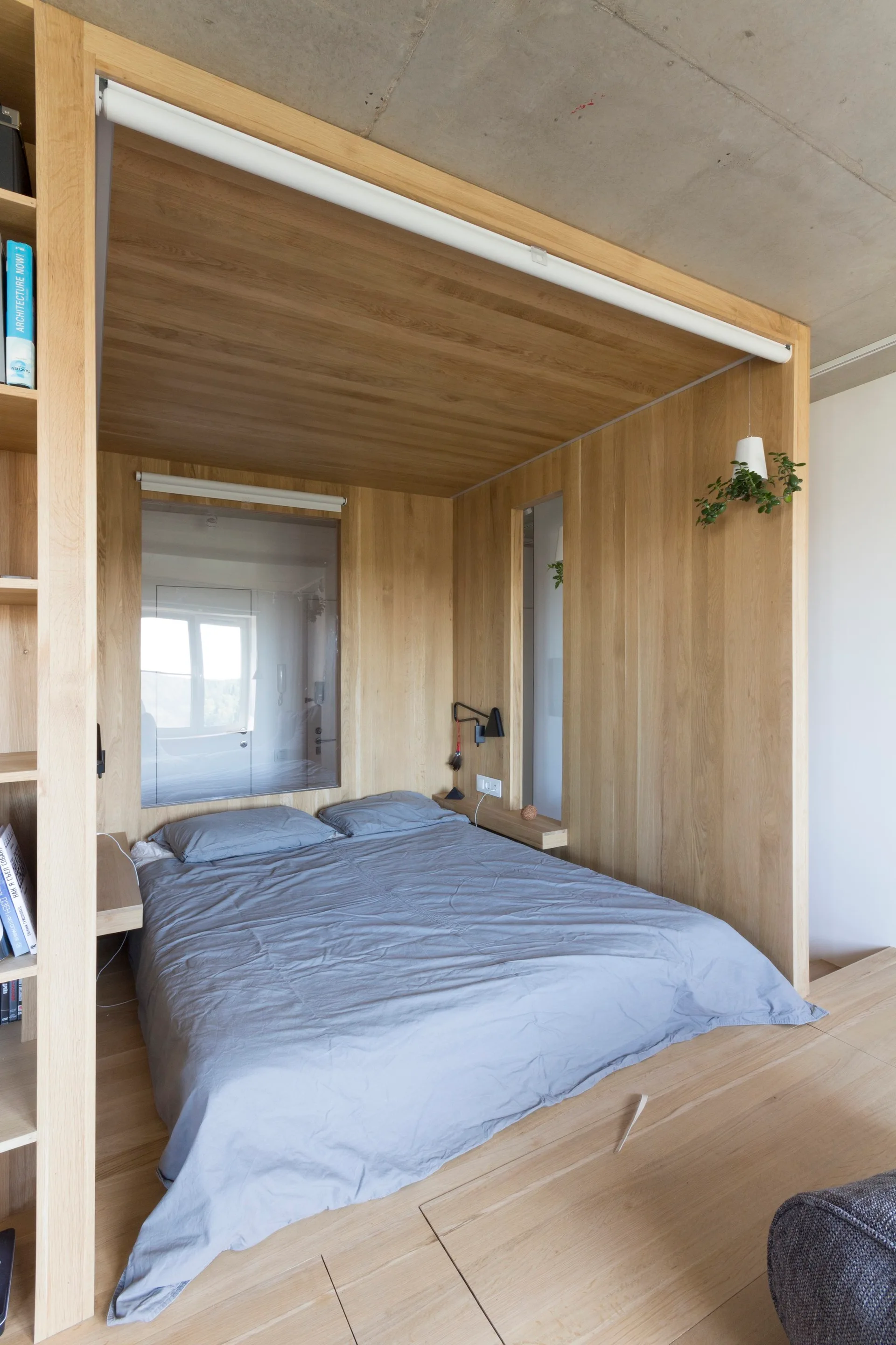A minimalist bed with grey linens is built into a warm wood-paneled alcove beneath an exposed concrete ceiling with integrated shelving.
