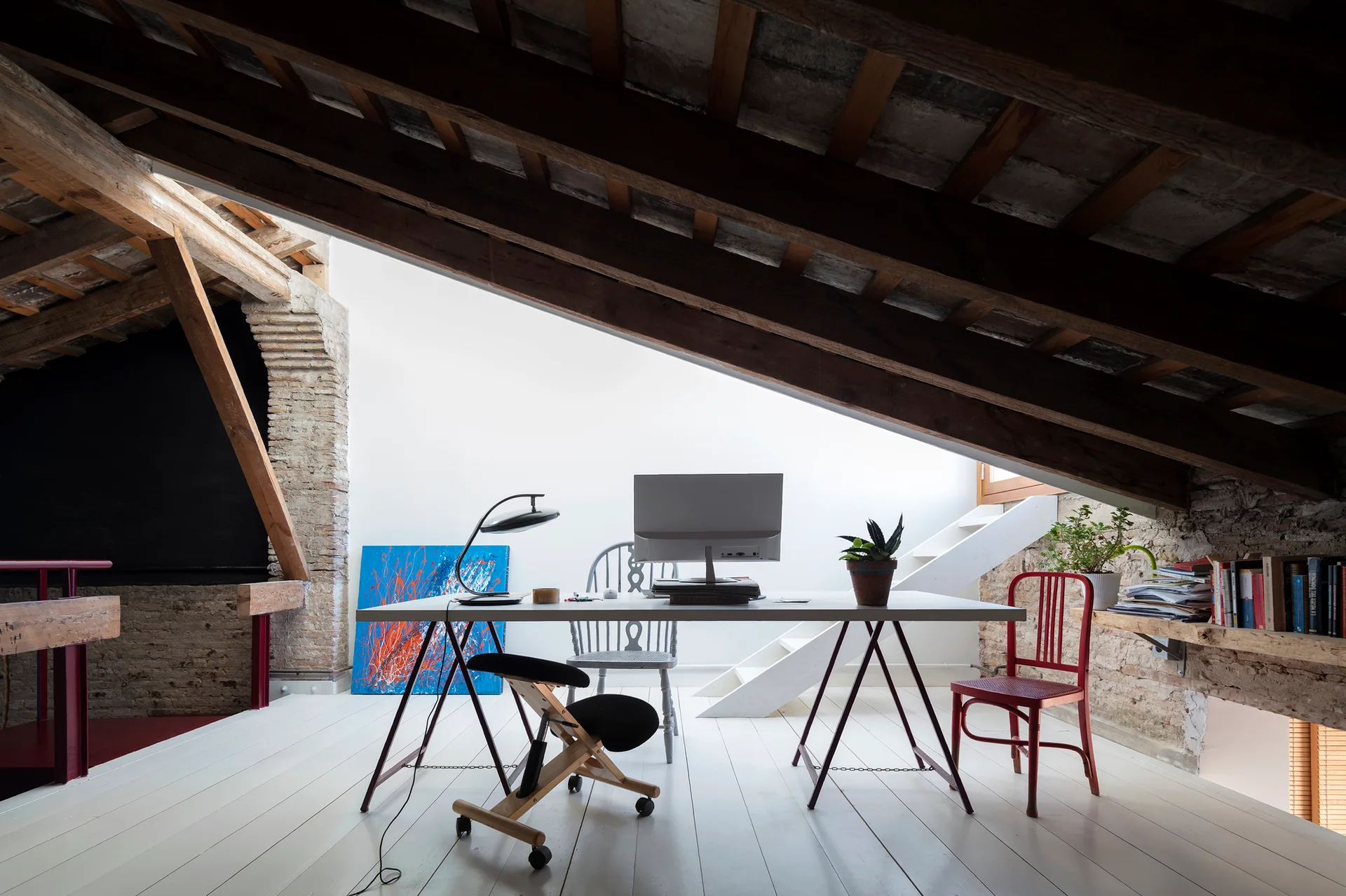 An attic office features a white trestle desk, computer monitor, and mismatched chairs under dark exposed wooden ceiling rafters.